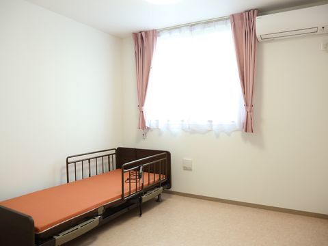 自然光の入る窓を設けたベージュの床に白い壁、ピンクのカーテンをつけたシンプルな居住スペース。介護用ベッド、エアコンも完備している。