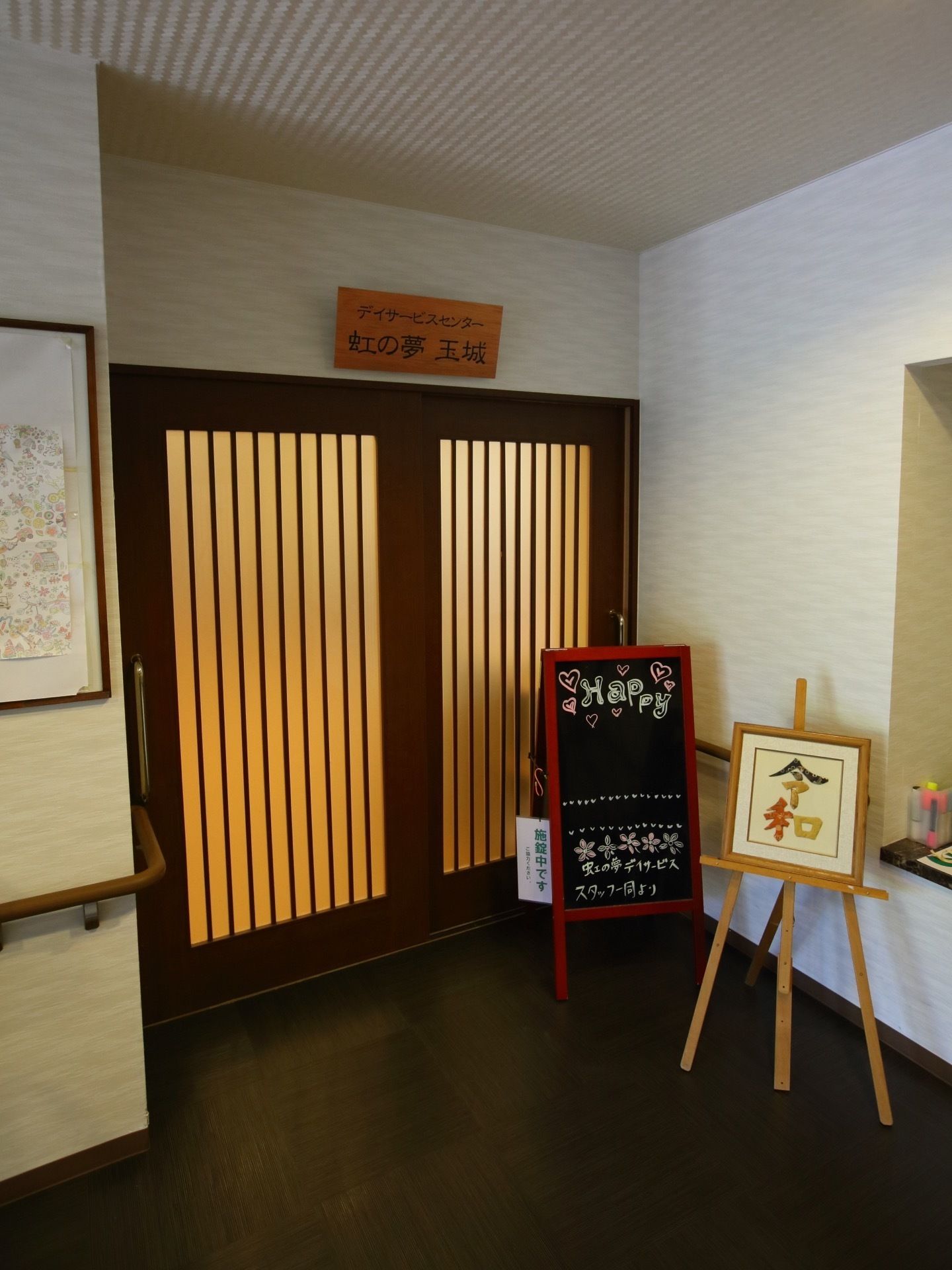 スッキリとした広いエントランスホールである。デイサービスセンターの扉は引き戸になっている。赤い縁のブラックボードが置かれている。