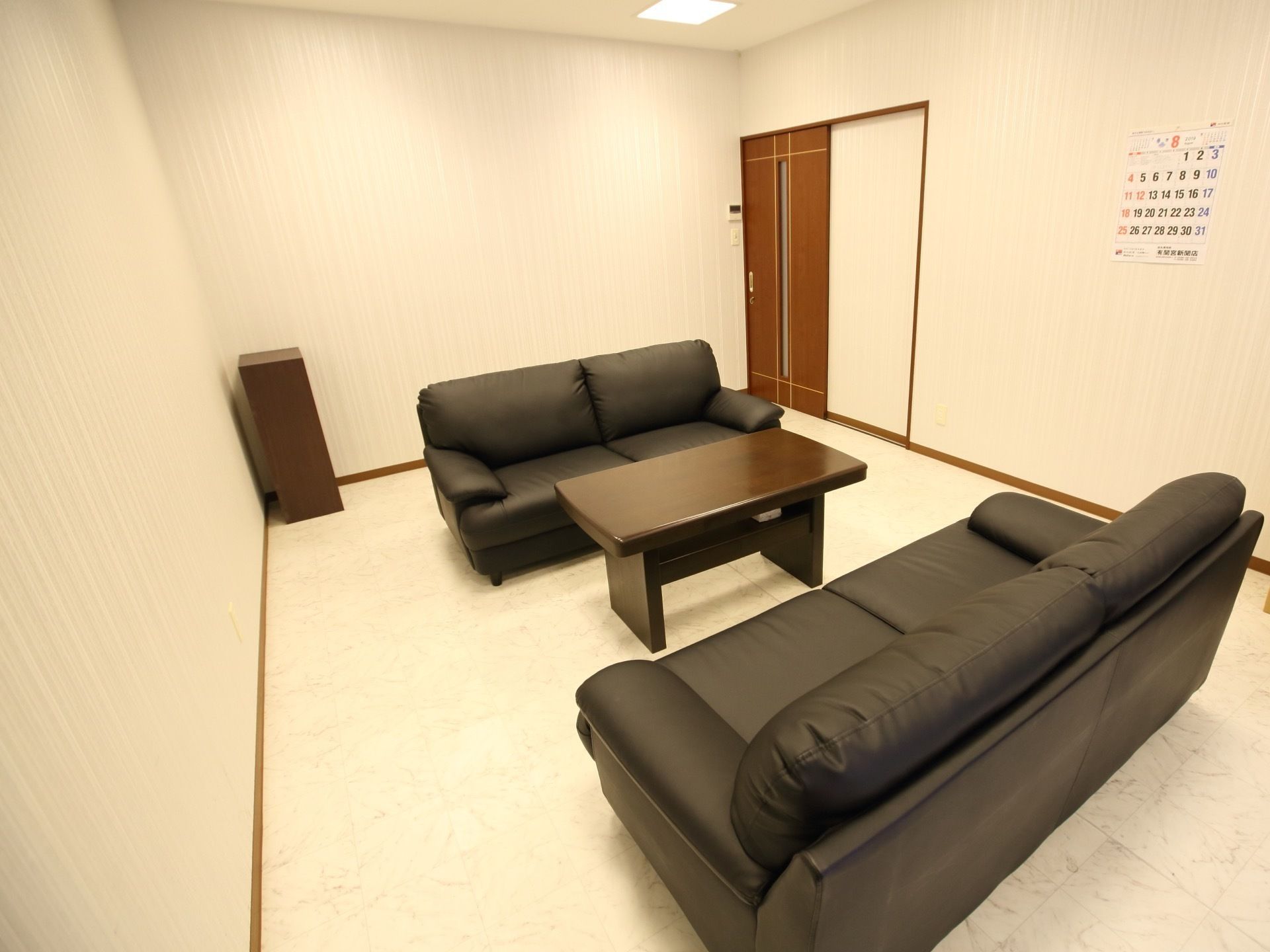 シンプルで落ち着いた雰囲気の相談室である。黒いソファとローテーブルが配置されている。壁にカレンダーが掛けられている