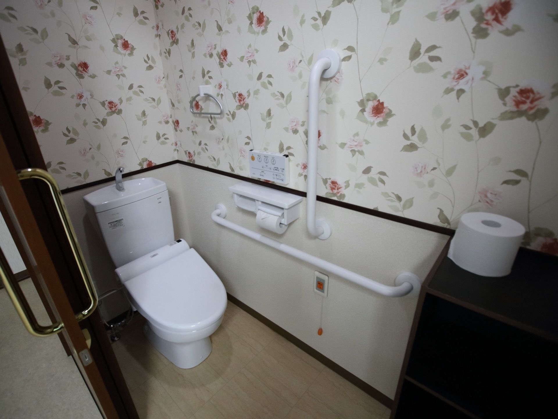 居室のトイレである。手すりや呼び出しボタン、温水洗浄便座が完備されている。カラーボックスが置かれている。