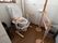 トイレの横には肘掛けが取りつけられている。隅にファンヒーターが置かれていて、その前には大きな手すりが設置されている。