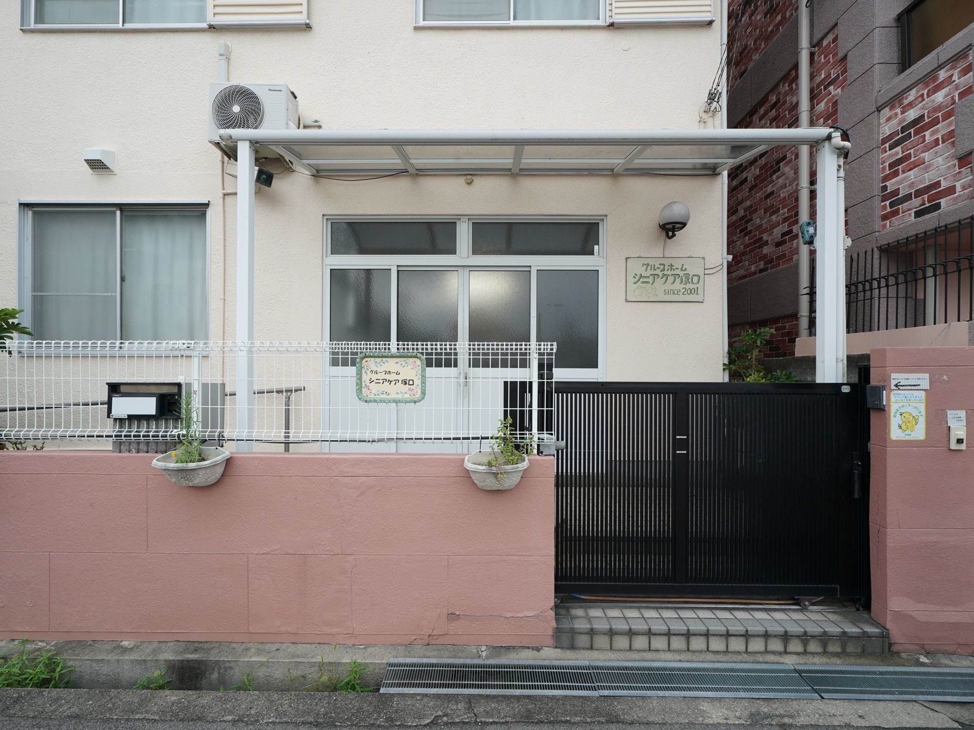 玄関は横開きの扉になっていて、屋根がついている。引き戸門扉がある。フェンスで囲まれており、鉢植えが飾られている。