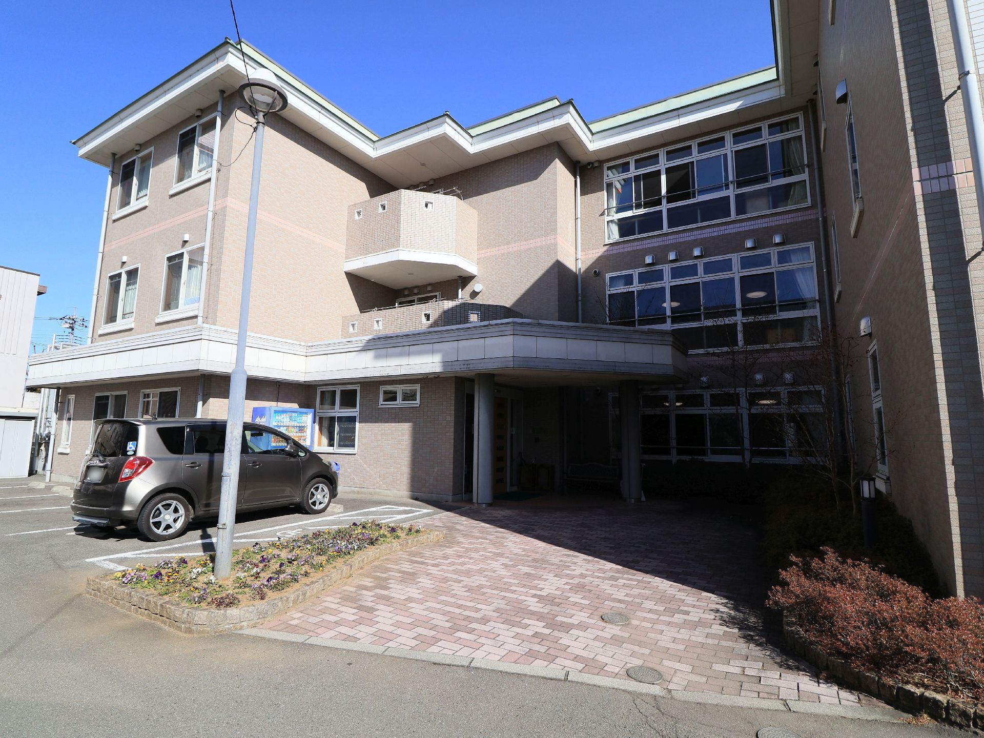 落ち着いた色合いの３階建ての建物になっている。施設前には駐車場が用意されており、車が１台停まっている。
