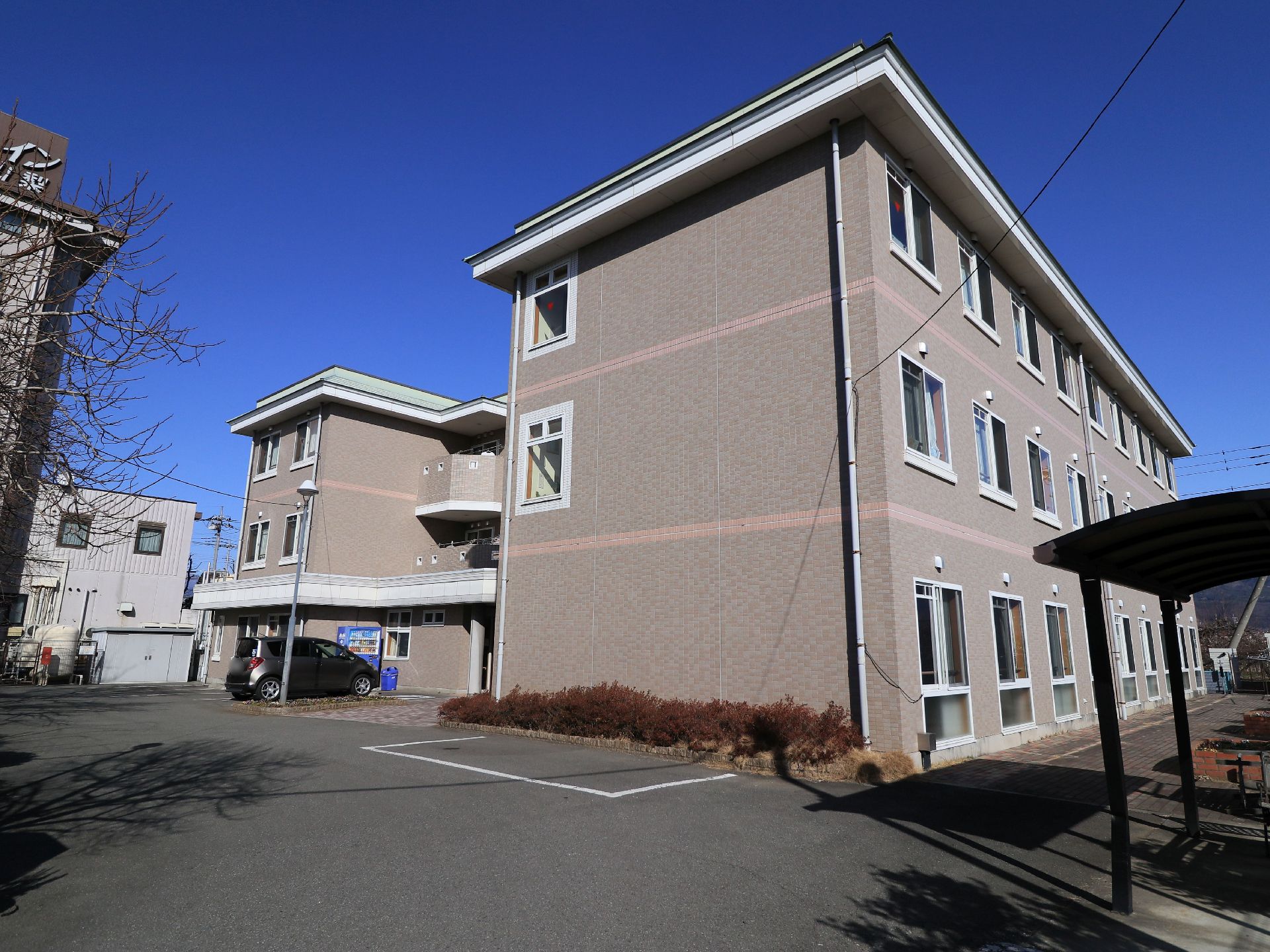 アットホームな雰囲気の３階建ての施設外観になっている。施設内には駐輪スペースや駐車場も用意されている。駐車場には車が１台停まっている。