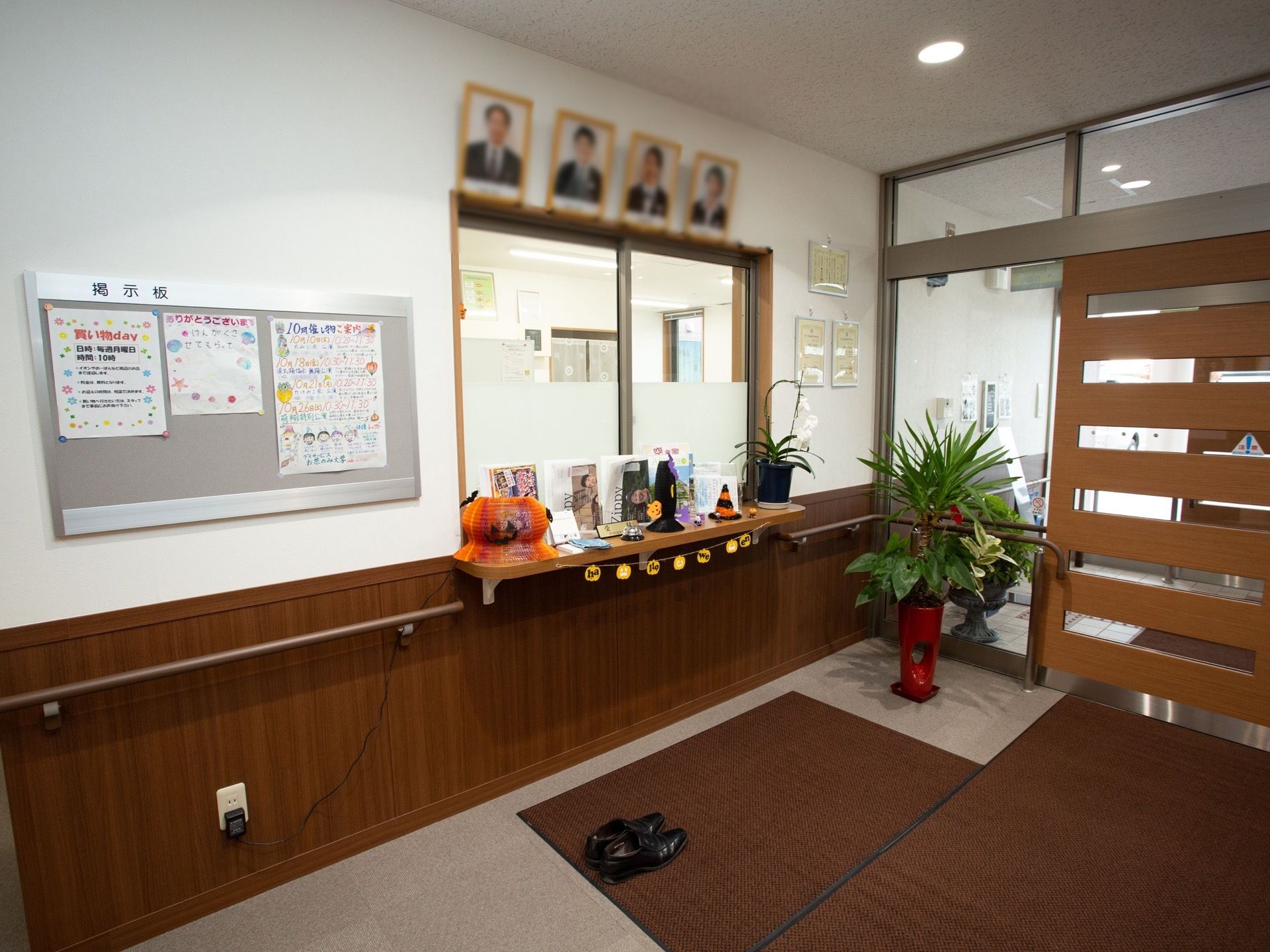 木目調のドアの手前の床に、複数の大判マットが敷いてある。左側の壁面に事務所の窓口があり、掲示板が設置されている。