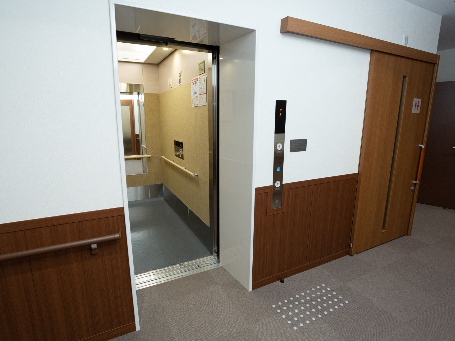 カーペット張りの通路の一角にエレベーターが設置されており、扉が開いている。隣にはトイレのドアが見える。