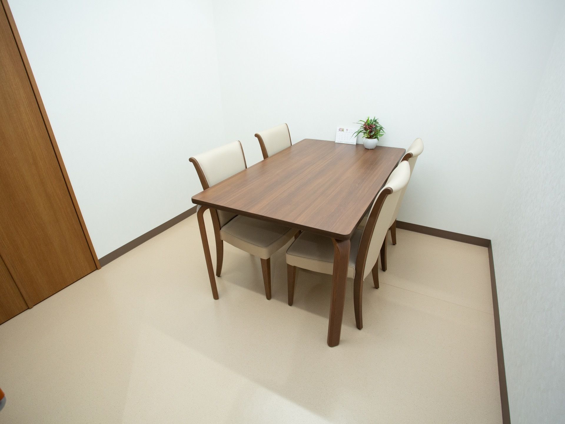 白い壁紙に囲まれた部屋の中央に、木目調のテーブルと白いクッションが施された椅子４脚が設置されている。テーブルの上に小さな観葉植物がある。