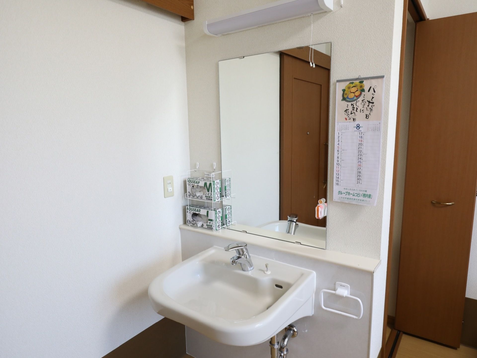 クローゼットの隣に設置されたシンプルな白を基調とした洗面台である。鏡の横にカレンダーが掛けられている。