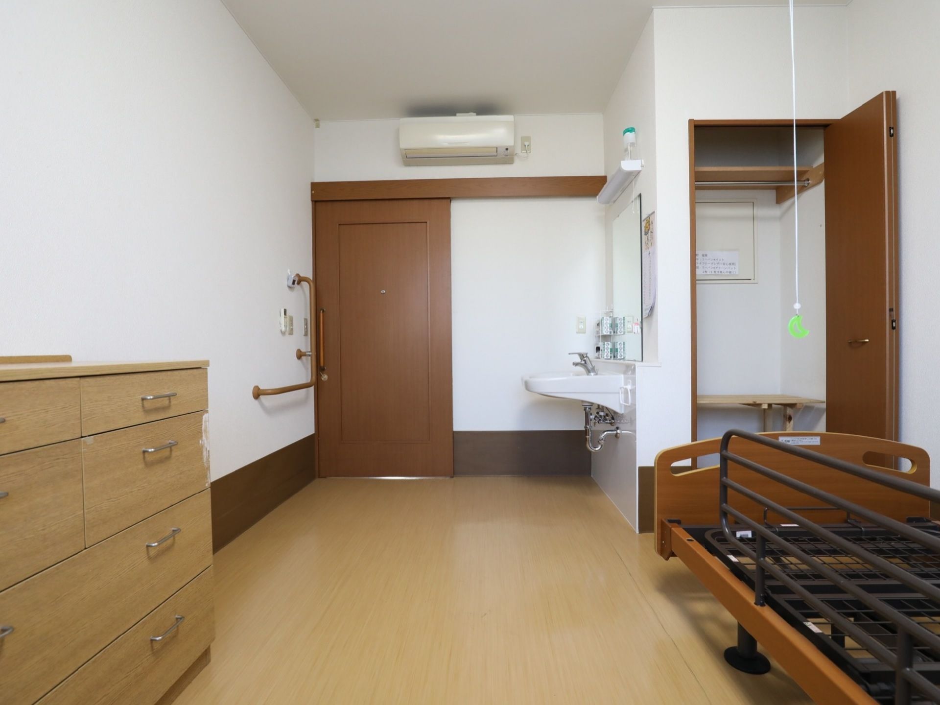 エアコンが完備された居室。洋服ダンスや介護ベッドが置かれており、入口横の壁にはＬ字型の手すりが設置されている。