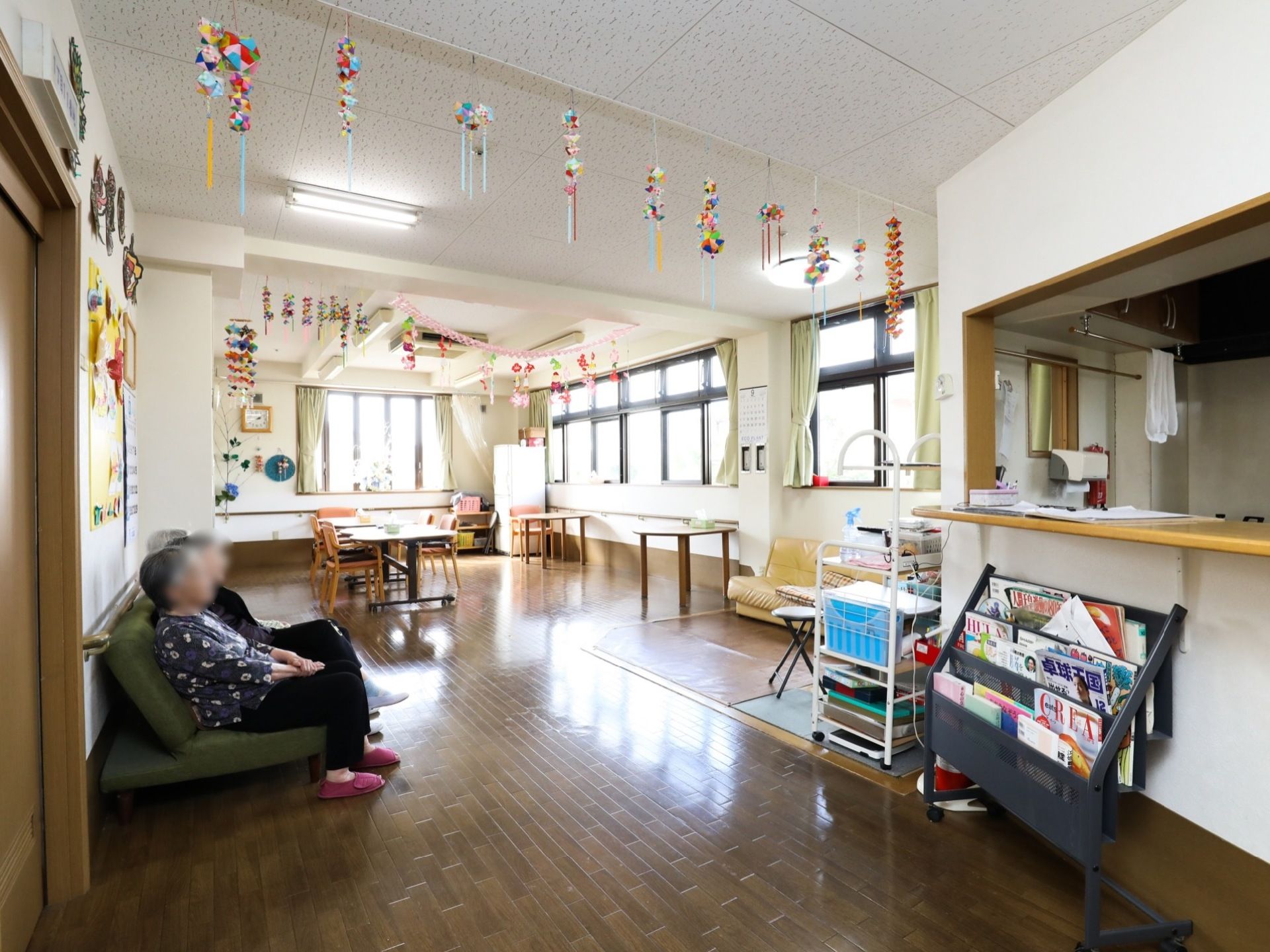 板張りの床に白い壁、天井の室内には、テーブルや椅子、ソファーや雑誌が置いてあり、入居者様がくつろいでいる。