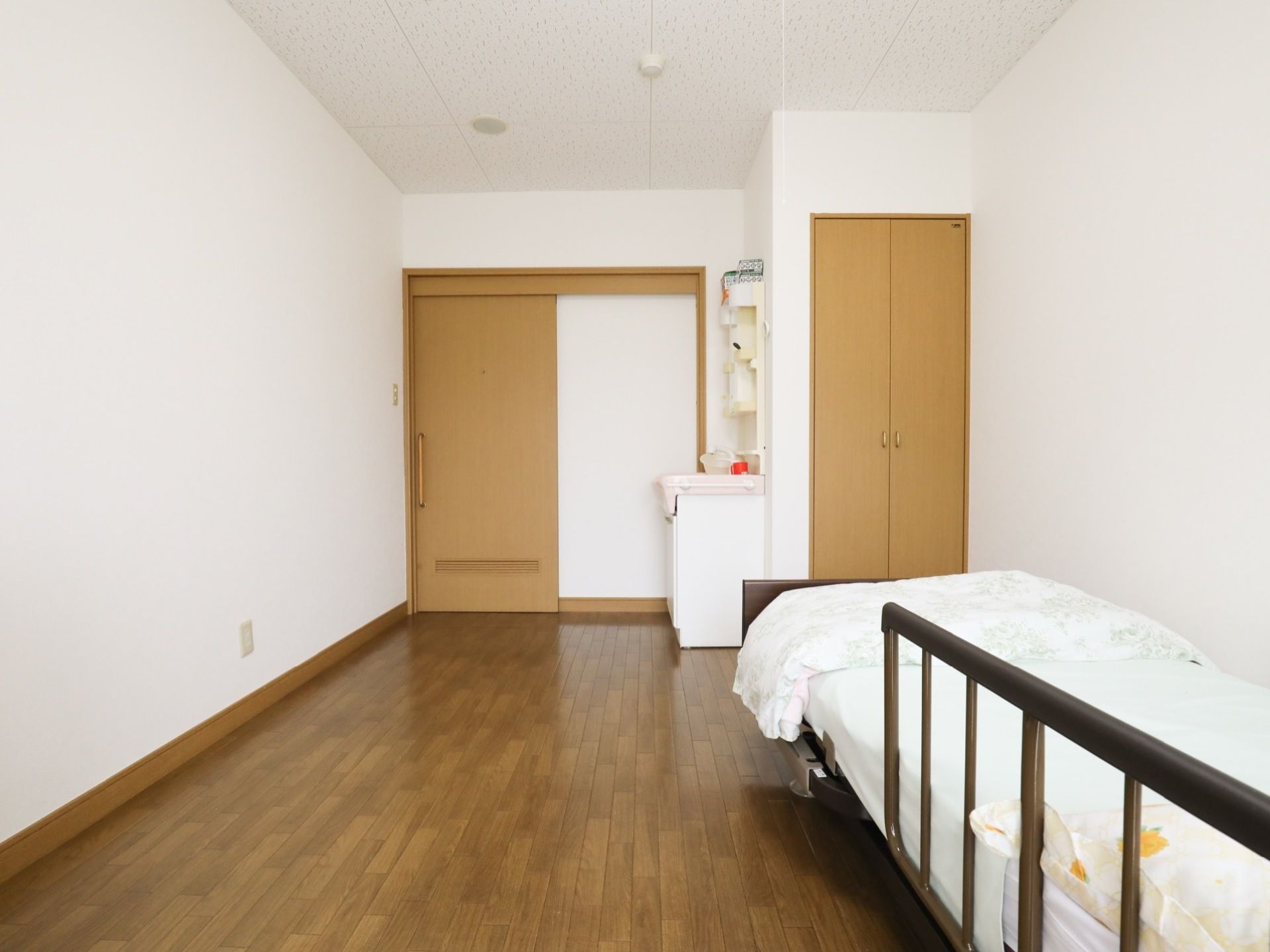 洗面台、ベッド、収納スペースがあるシンプルでコンパクトな居室である。ベッドは転落防止の柵が設置してあり、安全に配慮されている。