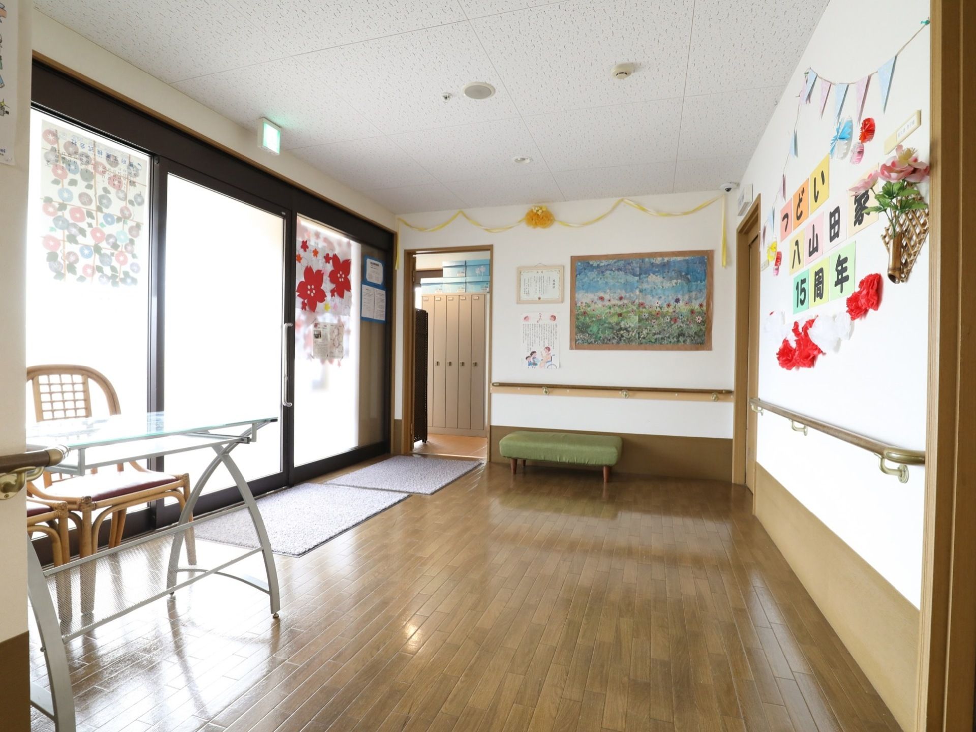 玄関ホールには、テーブルと椅子がガラス張りの壁面沿いに置いてある。白い壁面には15周年と表示された張り紙の周りに、貼り絵や花が飾られている。