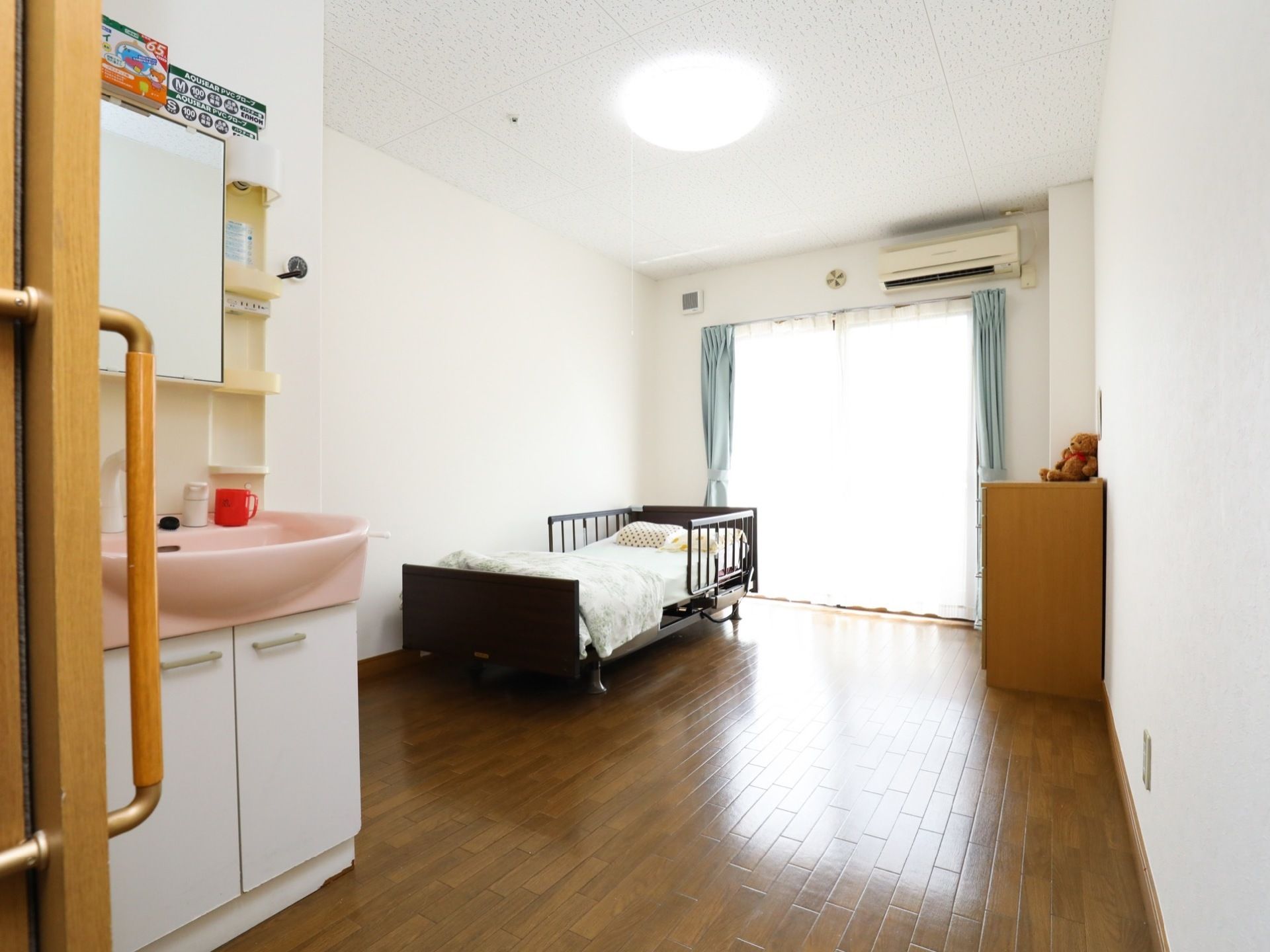 居室は、正面にガラス戸があり壁面上部にエアコンが設置されている。白い壁面沿いには介護用ベッドと向い側にはタンス、入口近くに洗面化粧台がある。