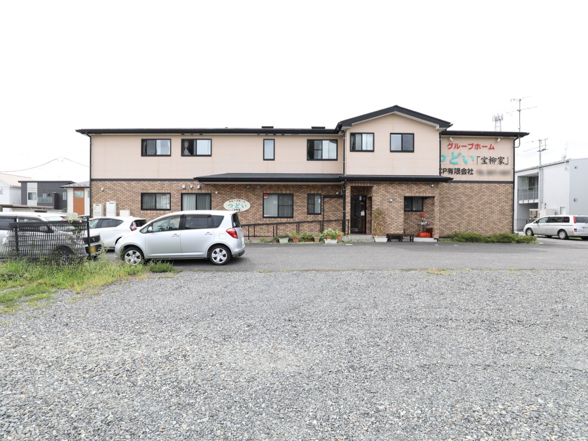 １階がレンガ調、２階が肌色の外壁の建物である。玄関周りに植物が並べられ、スロープが設置されている。駐車スペースが広く設けられている。