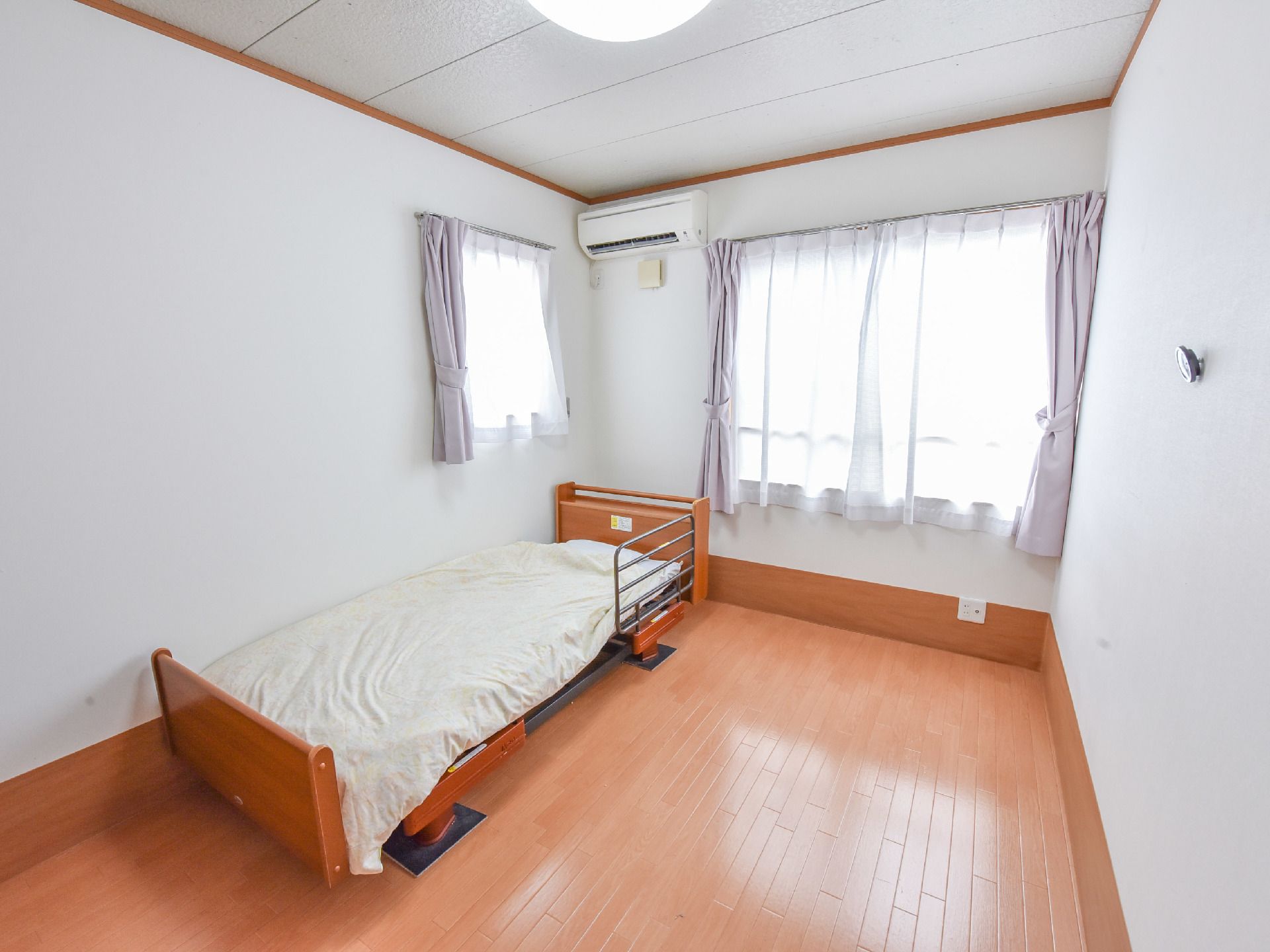 白色の天井と壁に茶色の床。正面には窓があり、横側にも一回り小さな窓がある。壁際には介護用のベッドが置かれている。