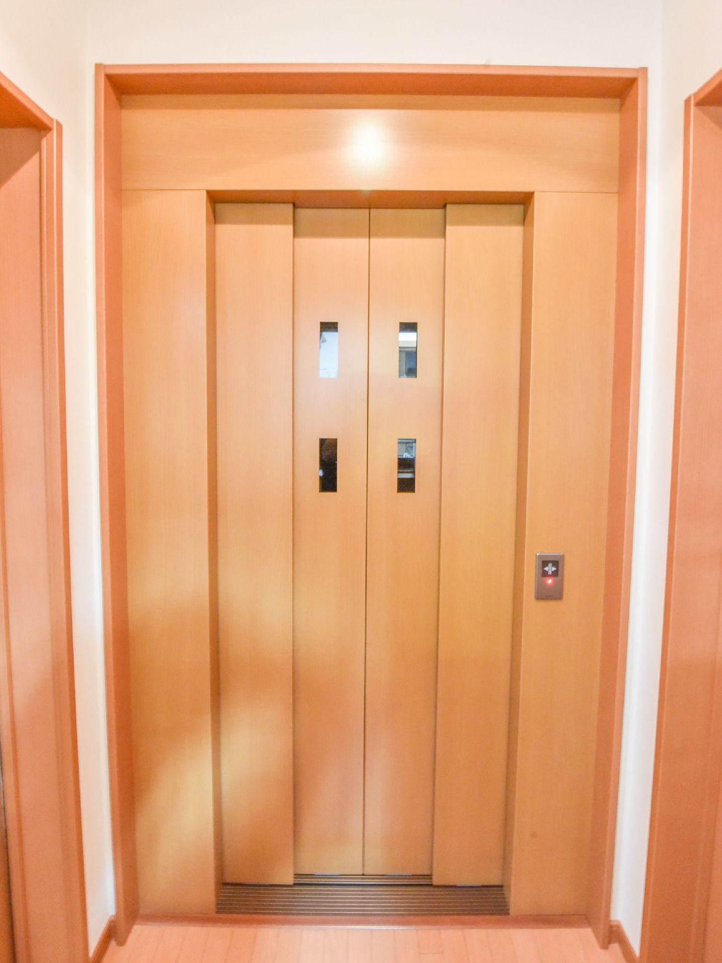 白色の壁に茶系のエレベータードア。小さな窓が4つ付いていて、内装の雰囲気に合わせて設計されたエレベータードアになっている。
