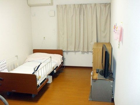 エアコンの下に、左右に手すりが取り付けられているベッドが置かれている。タンスの横にはテレビも置かれている。