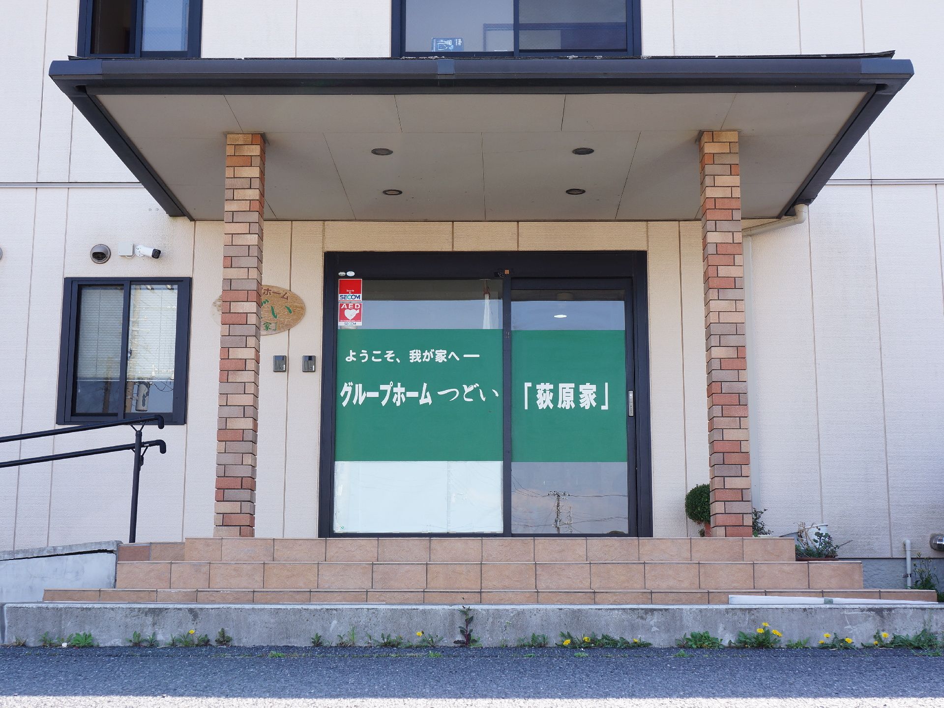 正面玄関の自動ドアには施設名と「ようこそ、我が家へ」のキャッチコピーが記されている。入居者は家庭的な環境の中で生活を送ることができる。