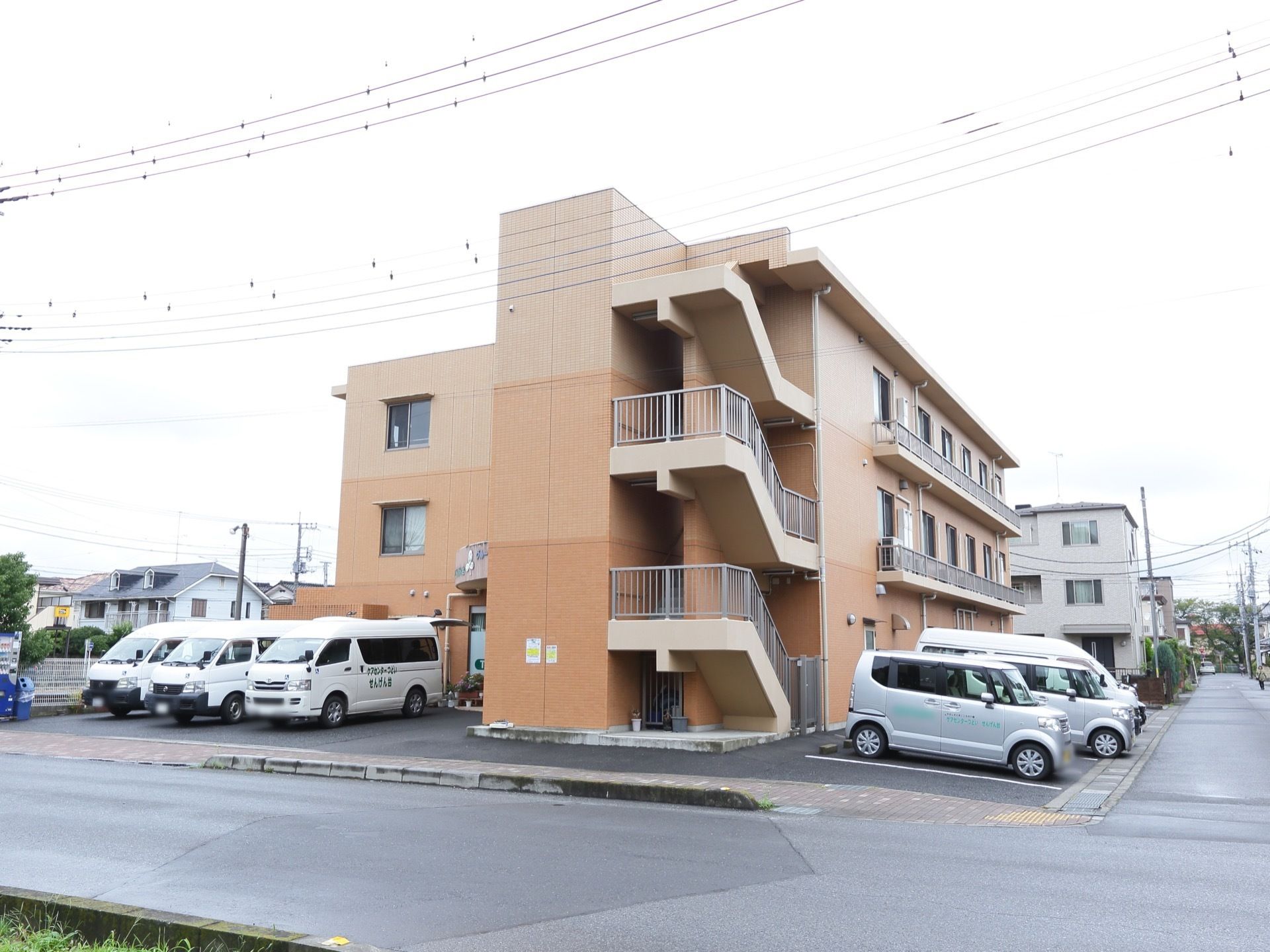 駐車場がある。自動販売機が置かれている。建物は3階建てとなっている。暖色で統一された優しい雰囲気の外観である。