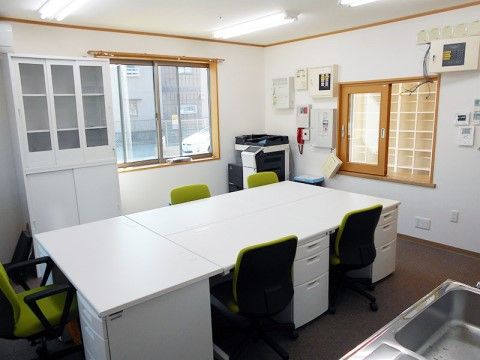 部屋の中央に、事務用の机や椅子が並ぶ。ミニキッチンやコピー複合機、本棚が設置されている。玄関とカウンター越しに会話ができる。