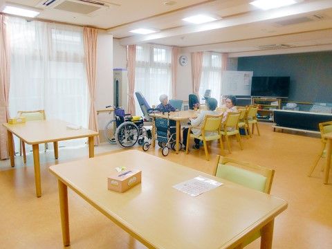 広々とした空間に、入居者様が１つの机を囲み談笑されている。壁際にはリハビリ機器やテレビが設置されている。