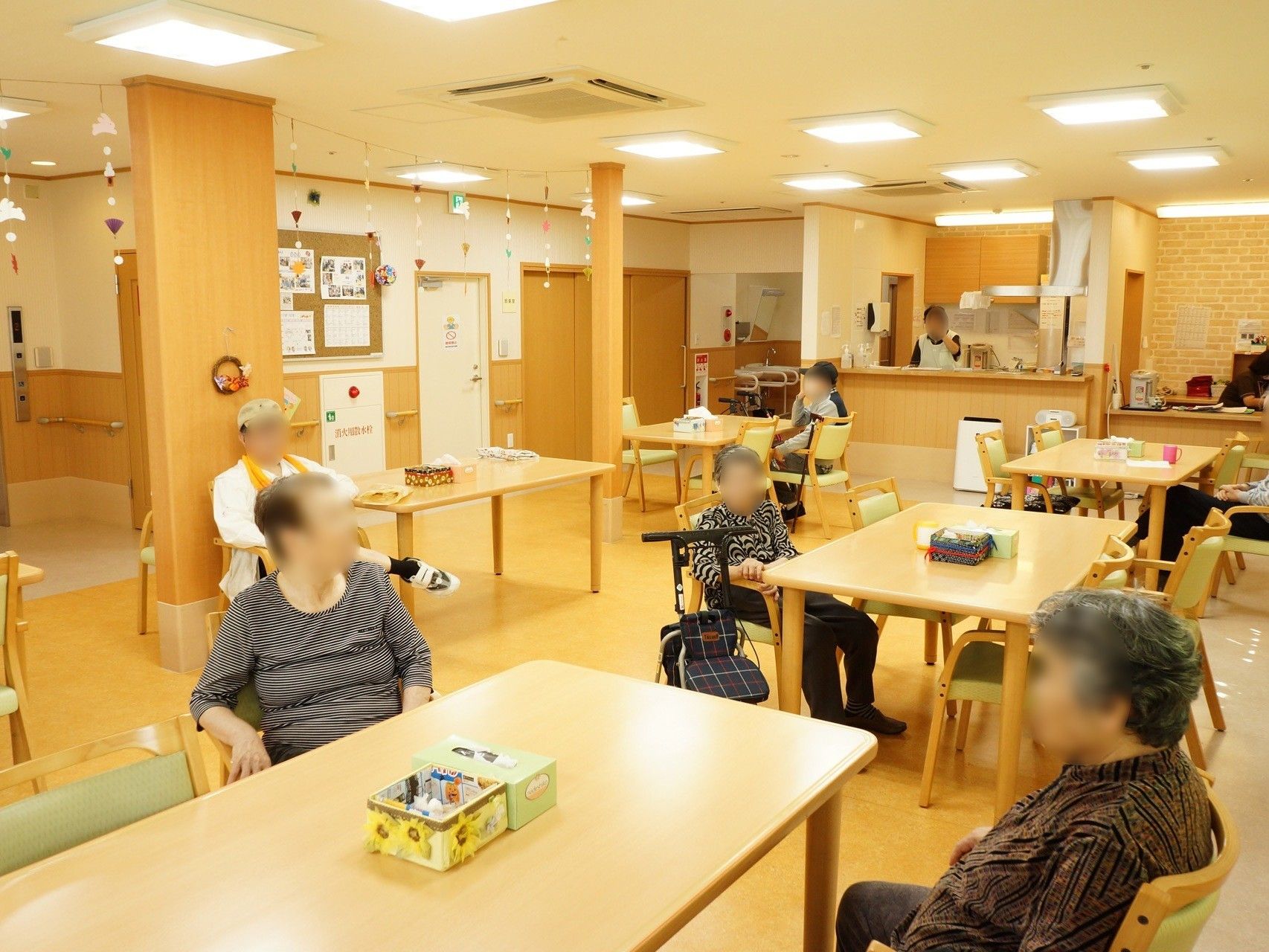 共有スペースには数人の入居者様がおり、お互い何かの話をしている様子となっている。奥にはスタッフがその様子を見つめている。