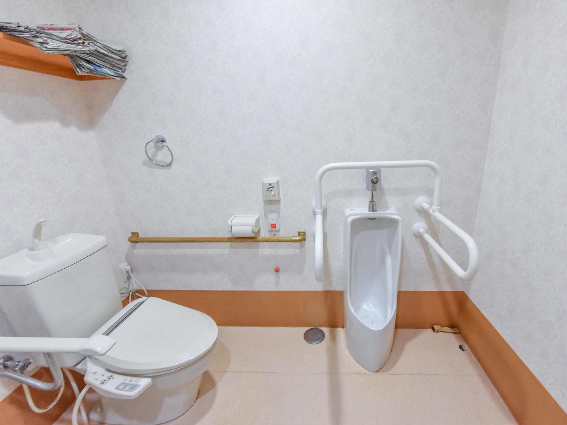 トイレは白色とベージュ色で統一されており、清潔感のある空間となっている。棚の上には、たくさんの新聞が積まれている。