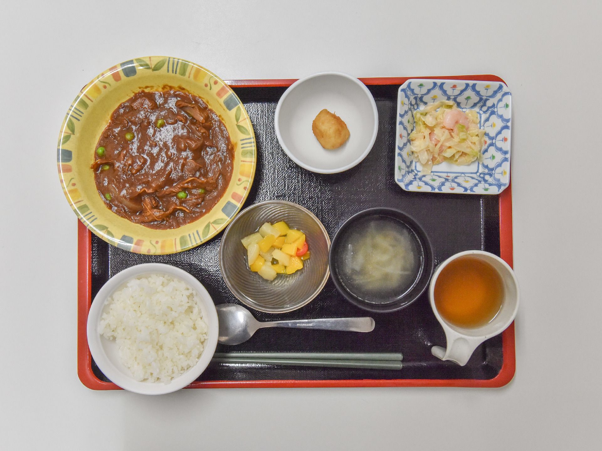 赤い縁の黒いトレーの上には、カレーや白米、スープや果物などの食事が並べられている。手前には、スプーンとお箸が置かれている。