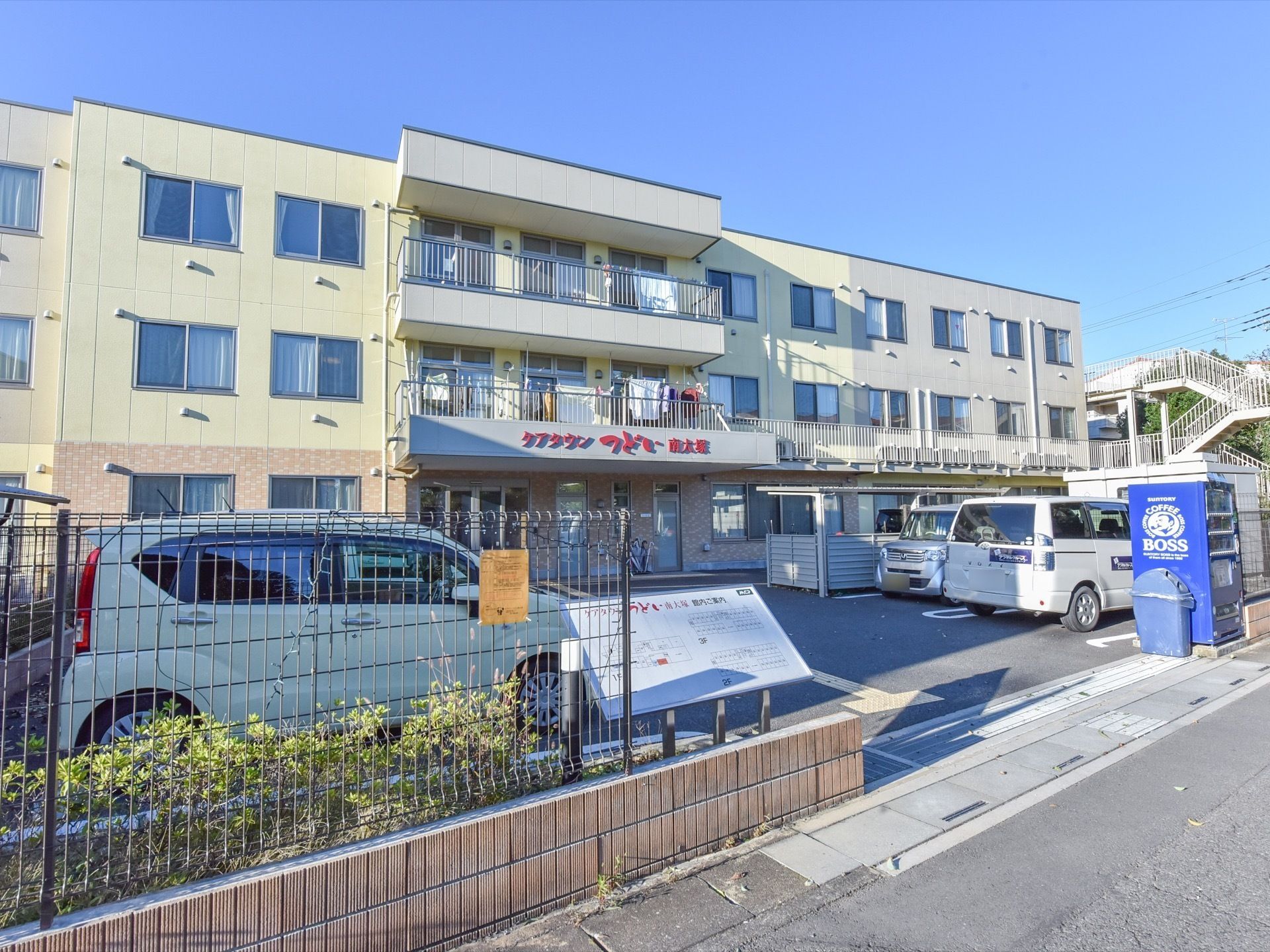 三階建ての施設である。玄関に大きく施設名が表示されている。一部にベランダが付いている。洗濯物が干してある。自動車が数台停車している。