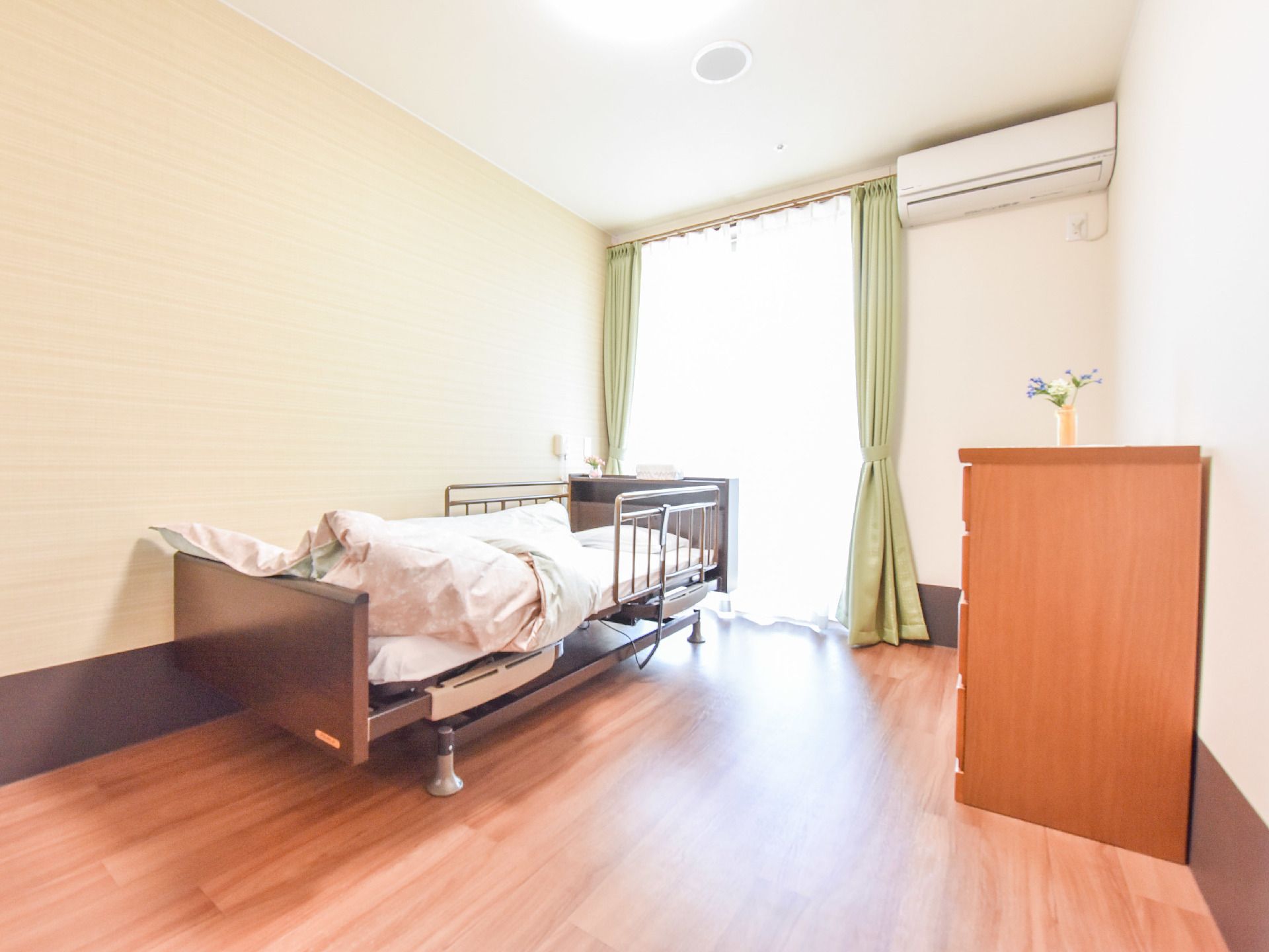 木目調の床に、天井まである掃き出し窓。窓の隣にエアコンがある。壁際に両手すり付きの低めのベッドがある。