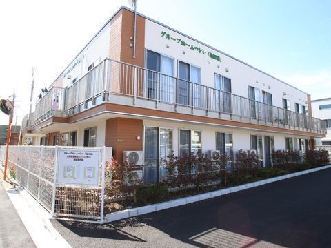 建物は1階部分は植栽帯で囲まれている。道路に面した部分は白い柵がある。2階には格子状手すりのバルコニーが建物沿ってある。