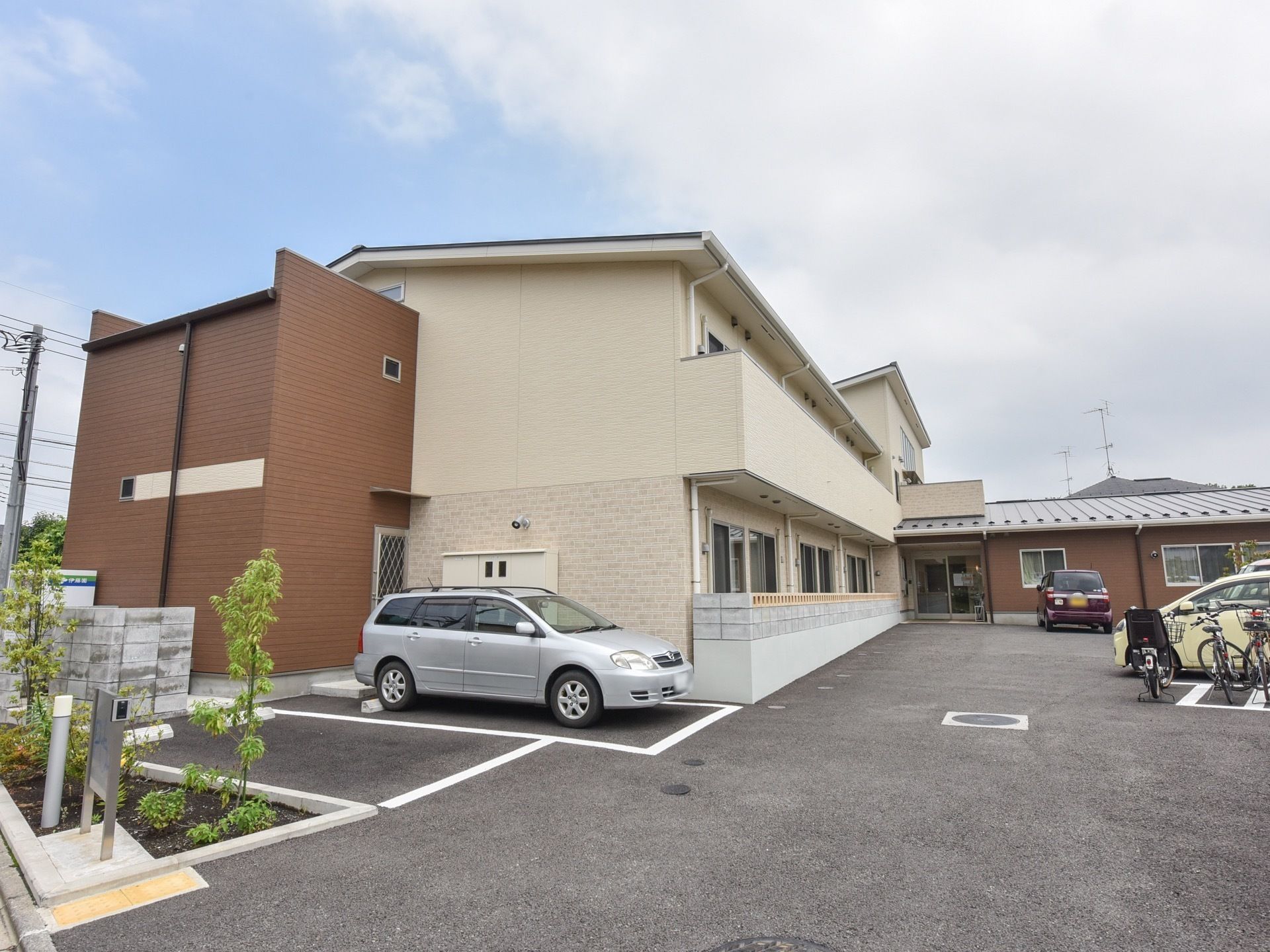 住宅街に違和感なく佇む、木造の２階建ての建物である。敷地内の駐車スペースには白線を引き、横側に緑の植栽を設けている。