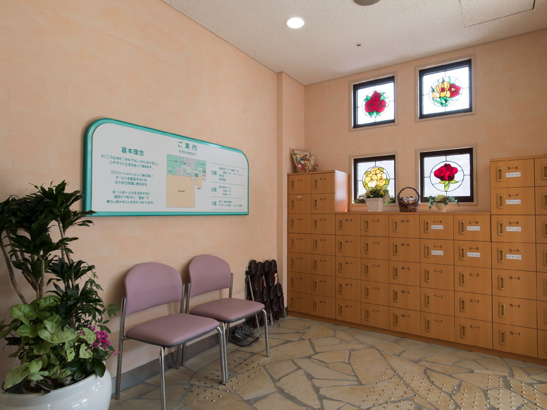 左側には観葉植物が置かれており、その右側には椅子が2脚並んでいる。中央には案内板がある。右側には靴箱がある。