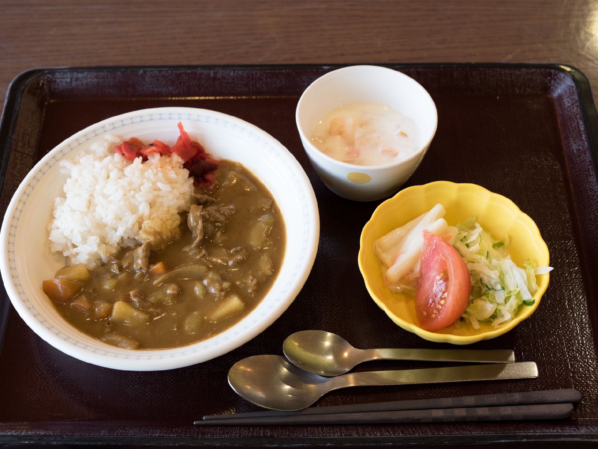 黒いお盆の上に様々なメニューが乗っている。左側にはカレーライスがある。右側にはサラダがあり、他に1品添えられている。