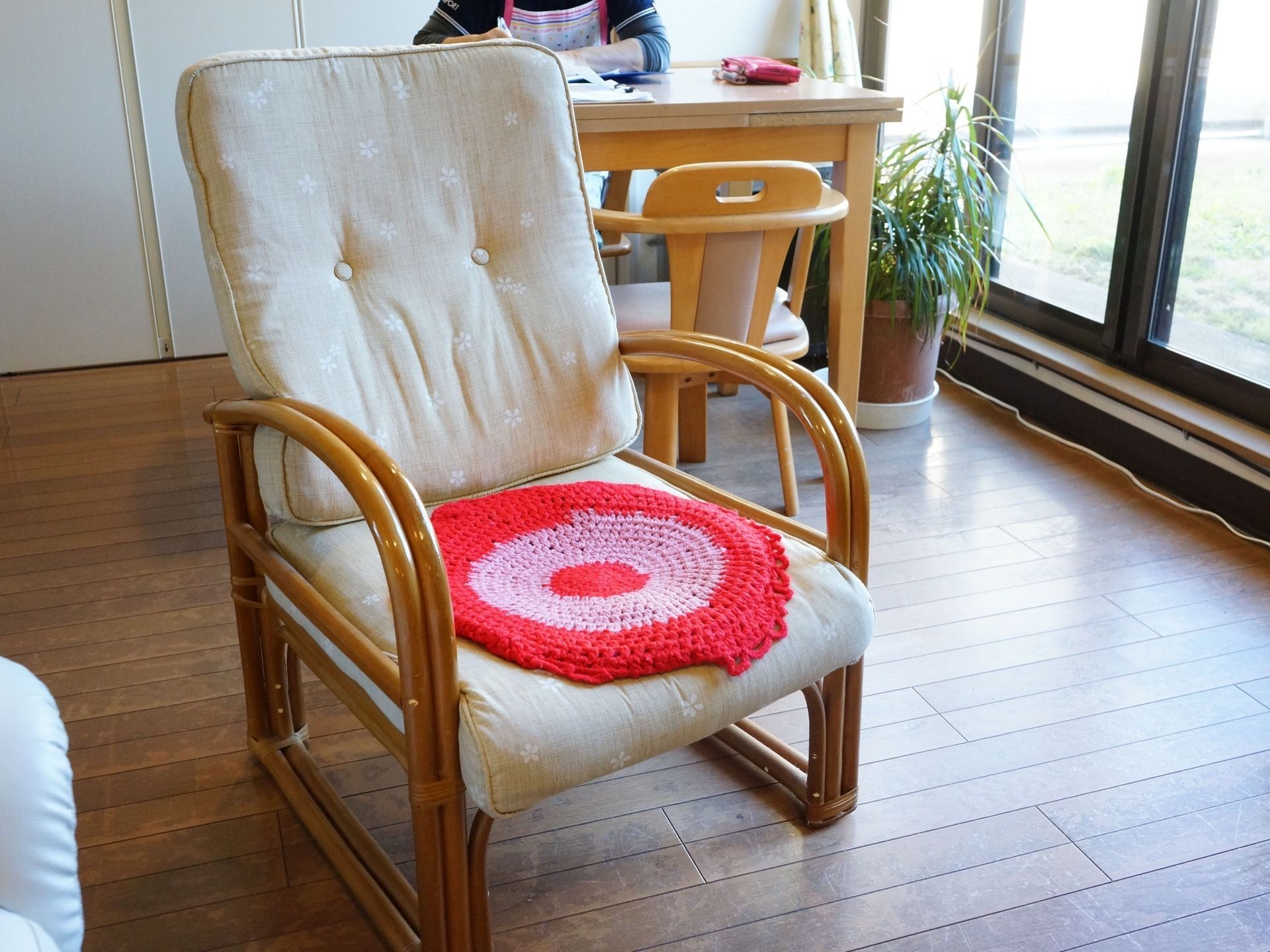 赤くて丸い座布団が置かれた椅子がある。その後ろでは、スタッフが机に座って事務作業を行っている。窓際に観葉植物が置かれている。