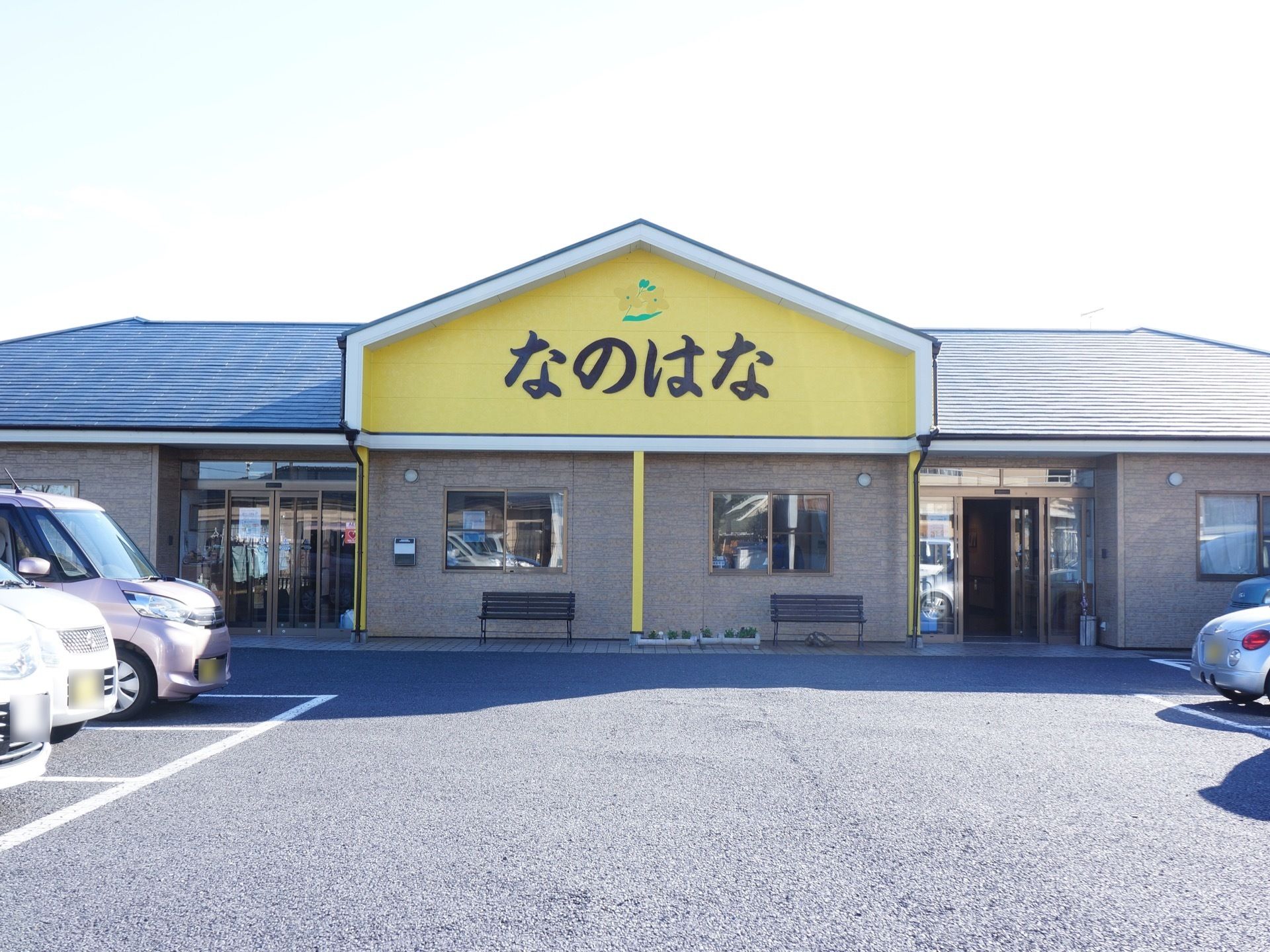施設の建物の正面にある黄色い壁面部分に大きな文字で施設名が記されている。敷地内には駐車スペースが確保されている。