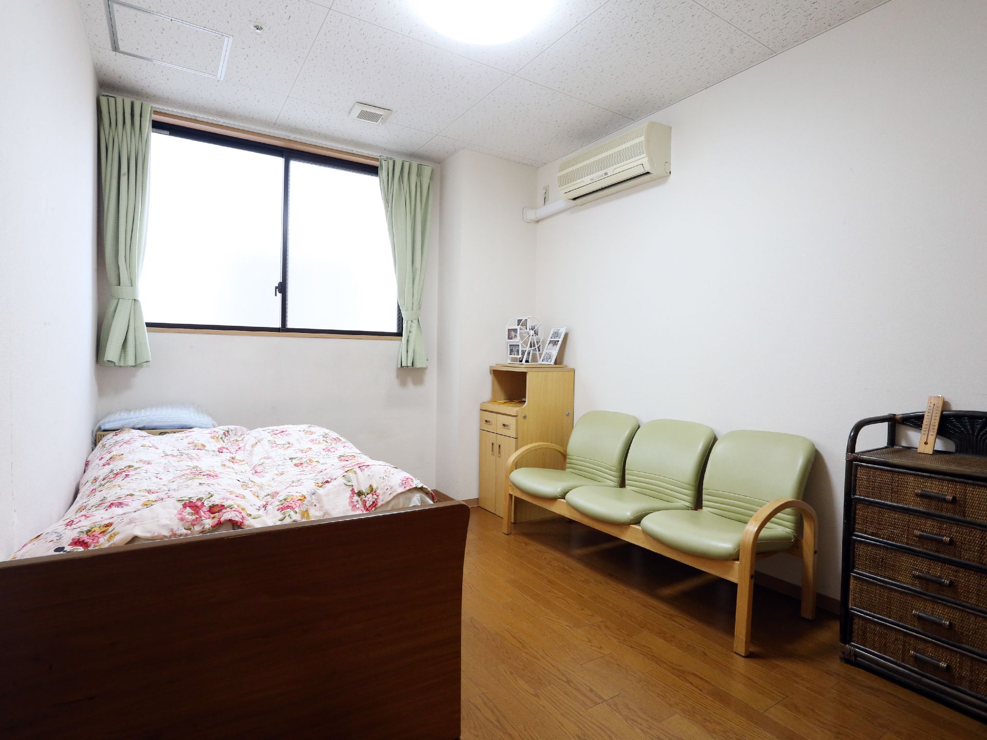 白基調の明るい居室である。窓には薄緑色のカーテンが設置されている。3人掛けのイスがある。右端の壁面上部にはエアコンが設置されている。