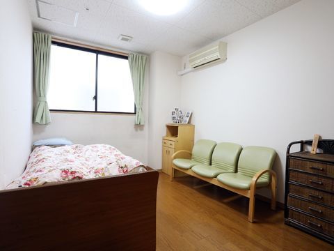 白基調の明るい居室である。窓には薄緑色のカーテンが設置されている。3人掛けのイスがある。右端の壁面上部にはエアコンが設置されている。