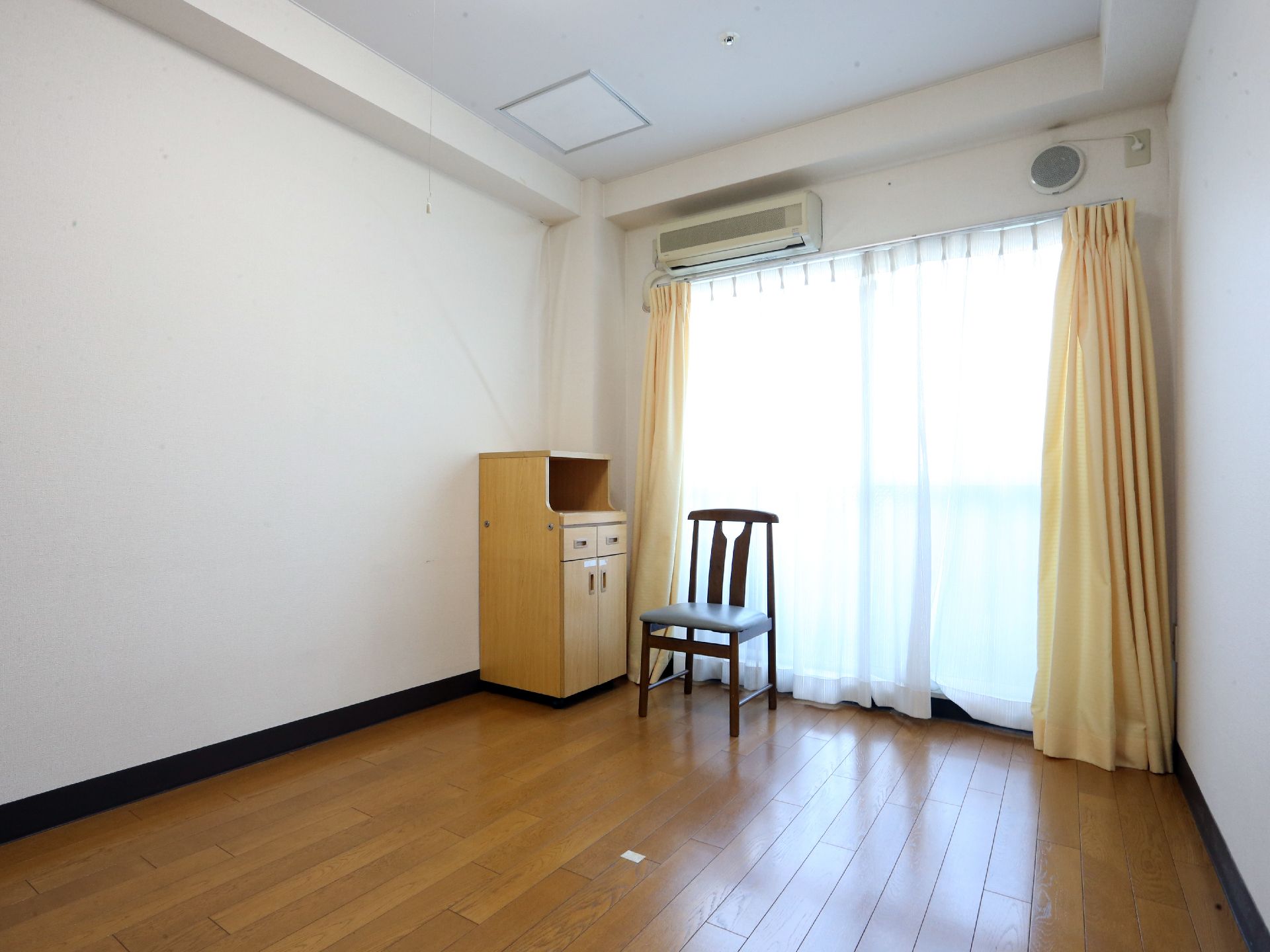 カーテン越しに外から明るい光が差し込んでいる。エアコンとイスと家具が設置されている。家具には引き出しが2つと開きが1つある。