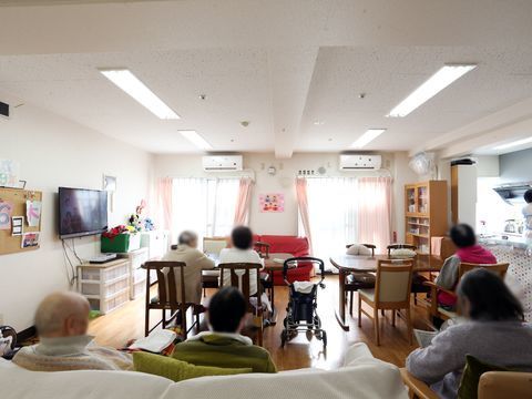 6名の入居者が座っている。エアコンが2台設置されている。2つある窓のあいだには紙細工のひな飾りがある。