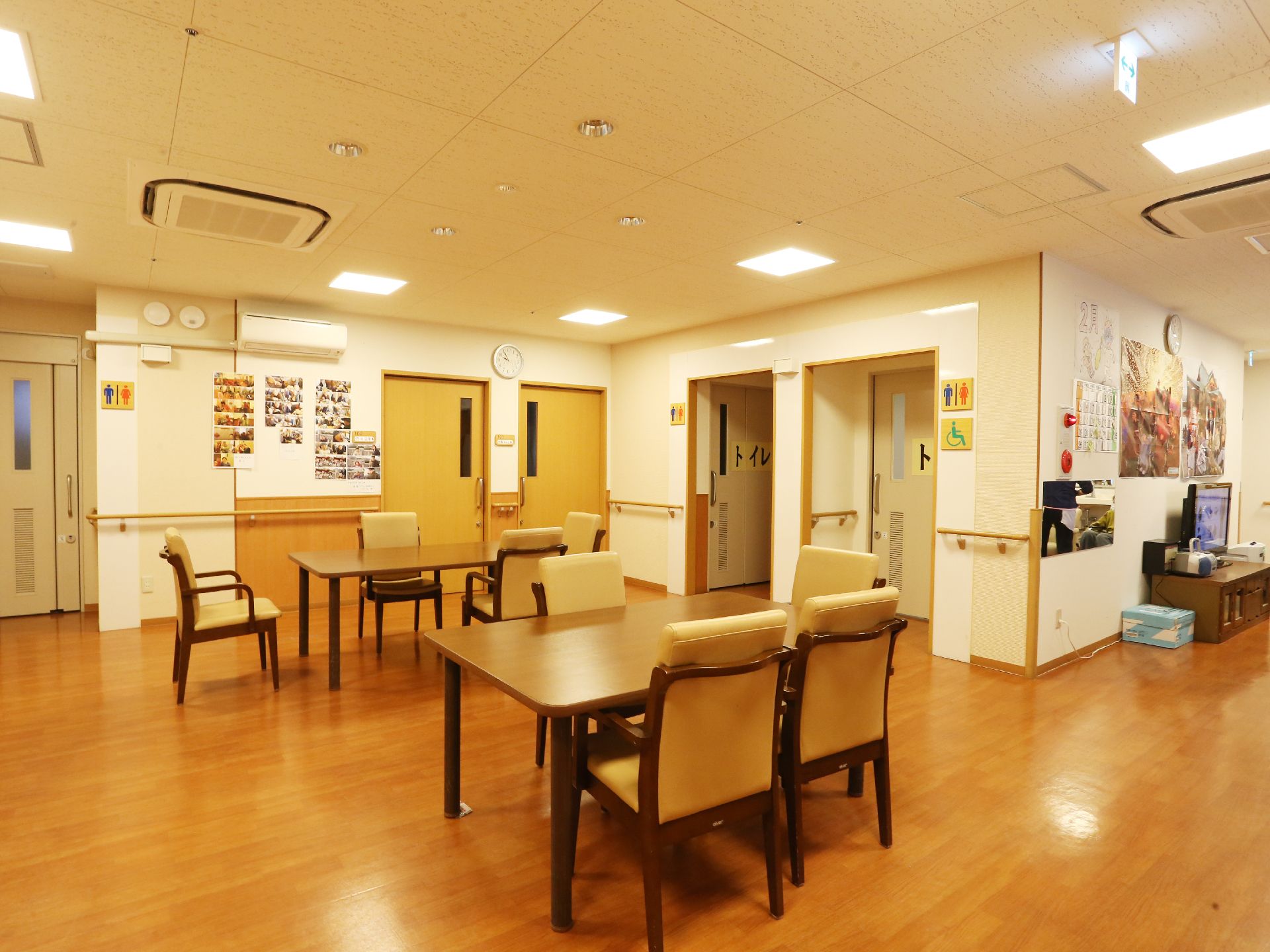 段差がなく、明るい木目調の床のお部屋。テーブルと椅子が複数おいてある。壁の各所には木製の手すりがあり、移動をスムーズに行える。