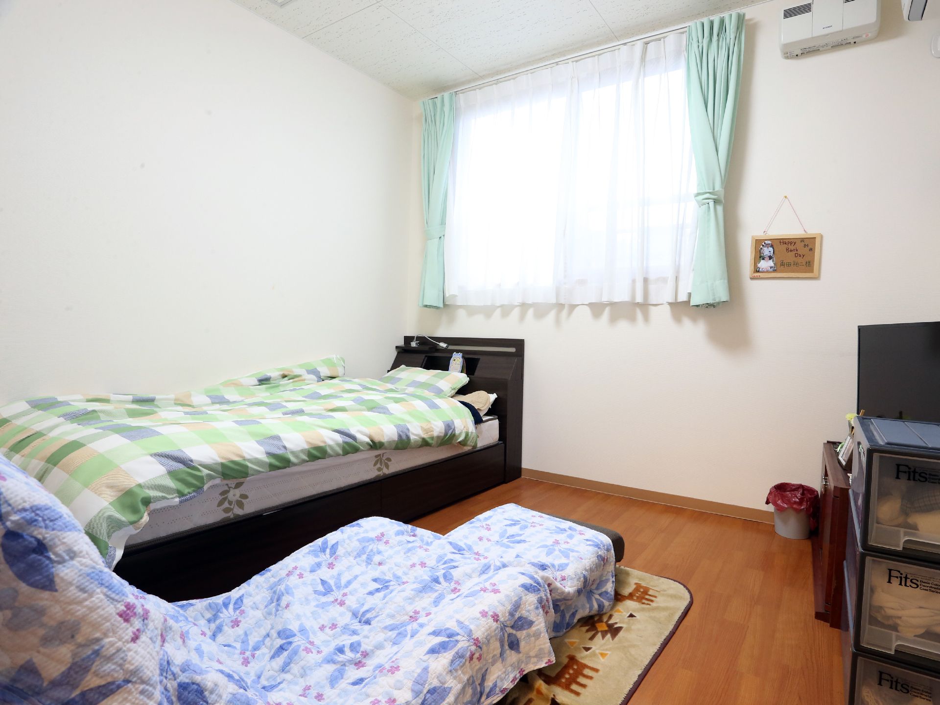 白い壁と、フローリングの部屋の窓際にベッドが置いてある。自然光が入り込み、くつろぎのある雰囲気である。