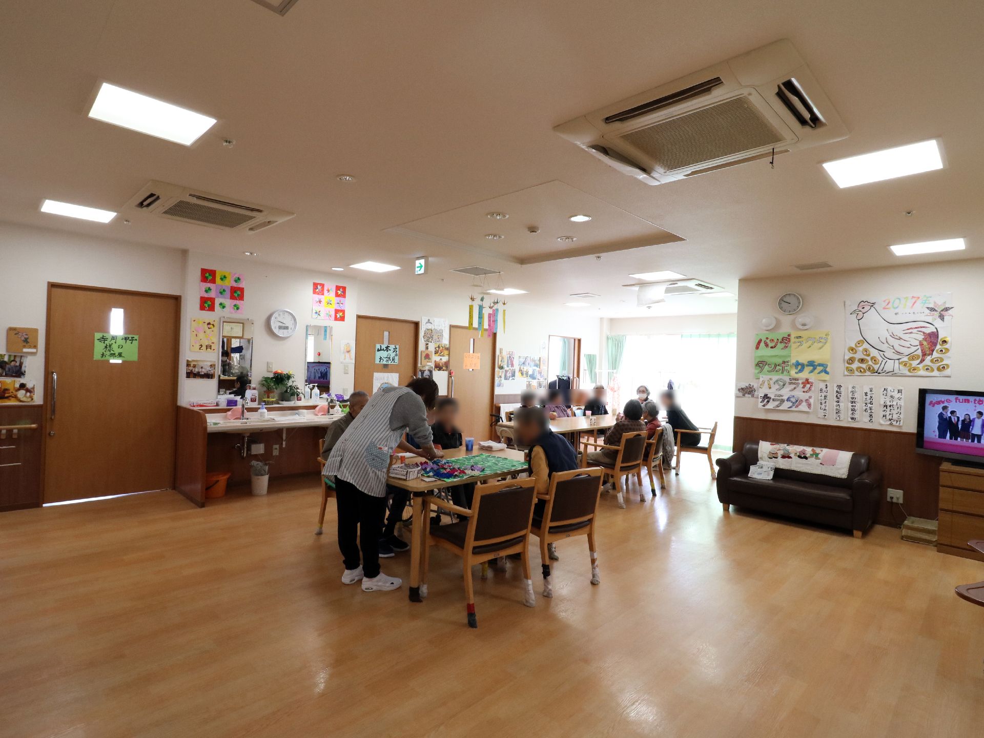 数人の入居者が、2つのテーブルに分かれて座っている。職員が1人いる。テレビがついている。壁面には様々な掲示物が貼付されている。