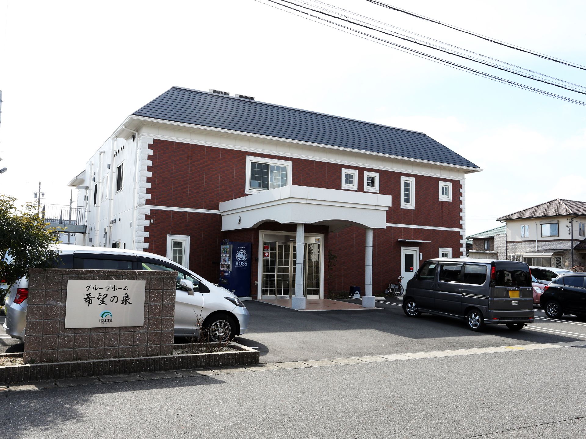 横の壁は白で、全面は茶色というデザインになっている。玄関横には自動販売機が置いてある。周囲には数台の車がとまっている。