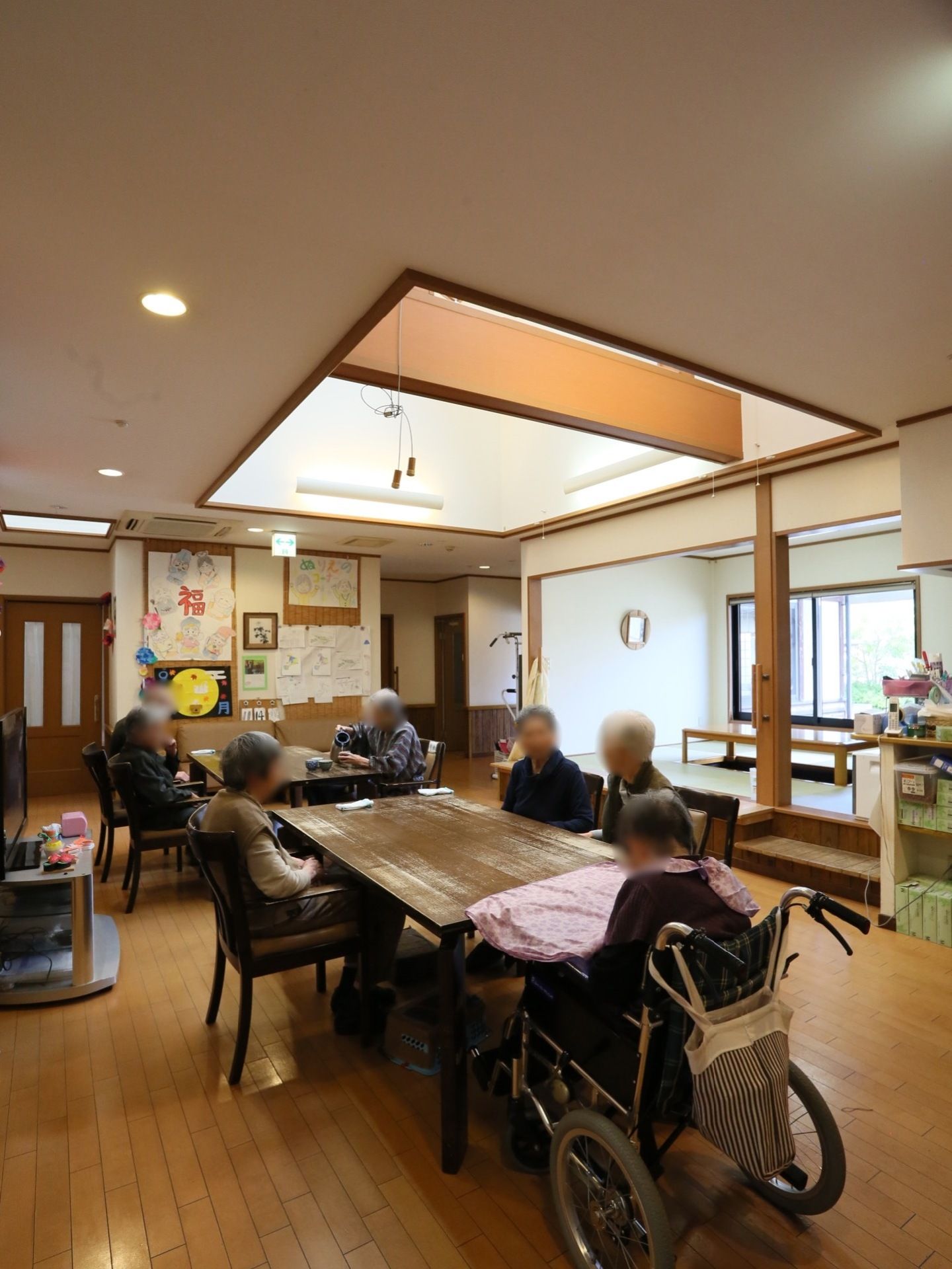 木製のテーブルが2つ設置されている。上部の天井には天窓が完備され光が注がれている。複数の入居者様がテーブルに着かれている。