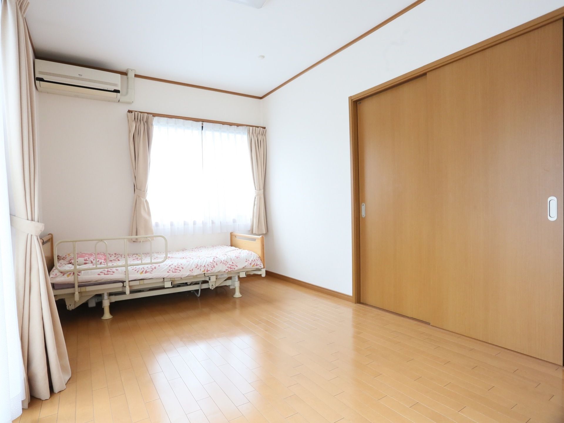 居室には、窓やエアコンが完備されている。木目調のフローリングに両開きの扉が設置され、介護用ベッドが置かれている。