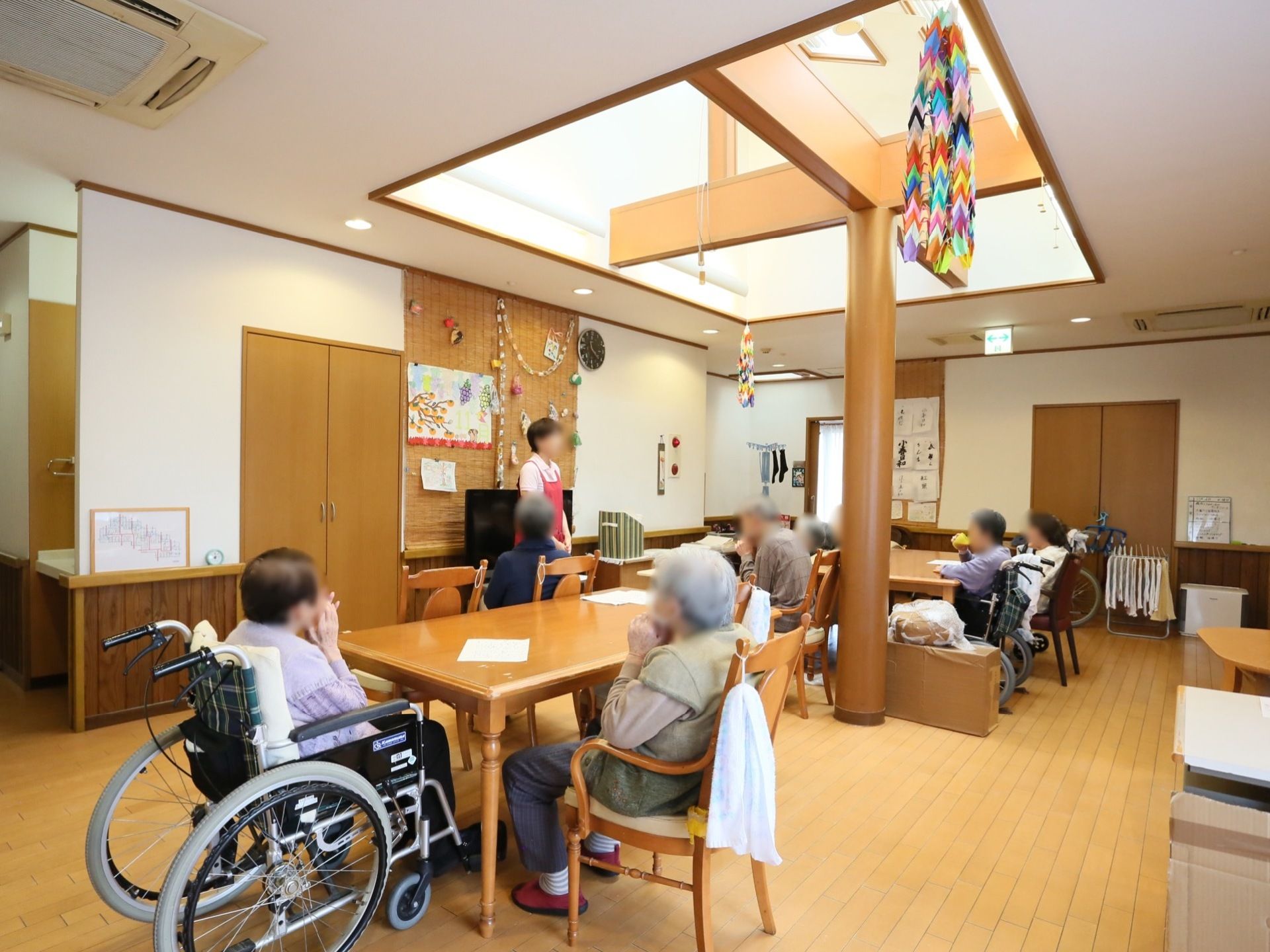共有スペースでは、椅子に腰掛けテーブルに着かれている入居者様、皆さまに向かい、1名の女性スタッフが前に立ち話をしている。