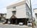 道路に面して３階建ての施設が建っている。建物の１階部分が駐車場になっており、複数の自動車が停まっている。