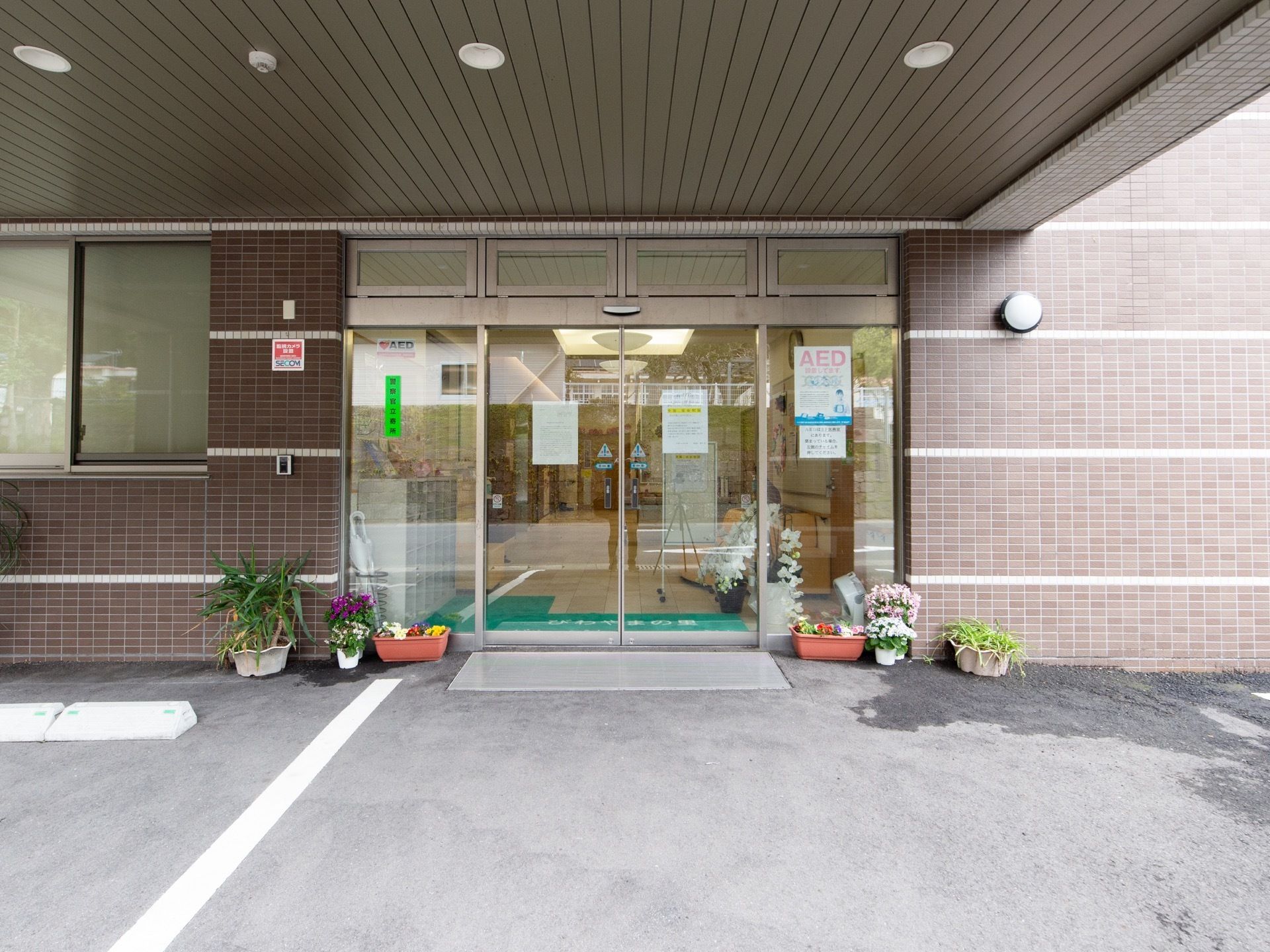 自動ドアの玄関の上部に、埋め込み式の丸型のライトが取りつけられた大きなひさしがある。ドアの前のプランターが置かれている。