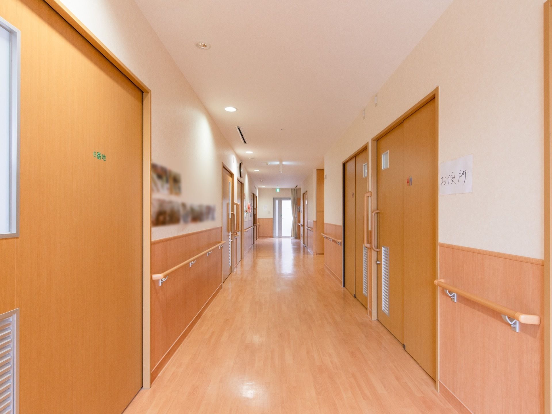 木目調の床の明るい廊下がある。ライトブラウンのドアが並んでおり、壁の半分は木の素材でできている。壁には手すりがある。